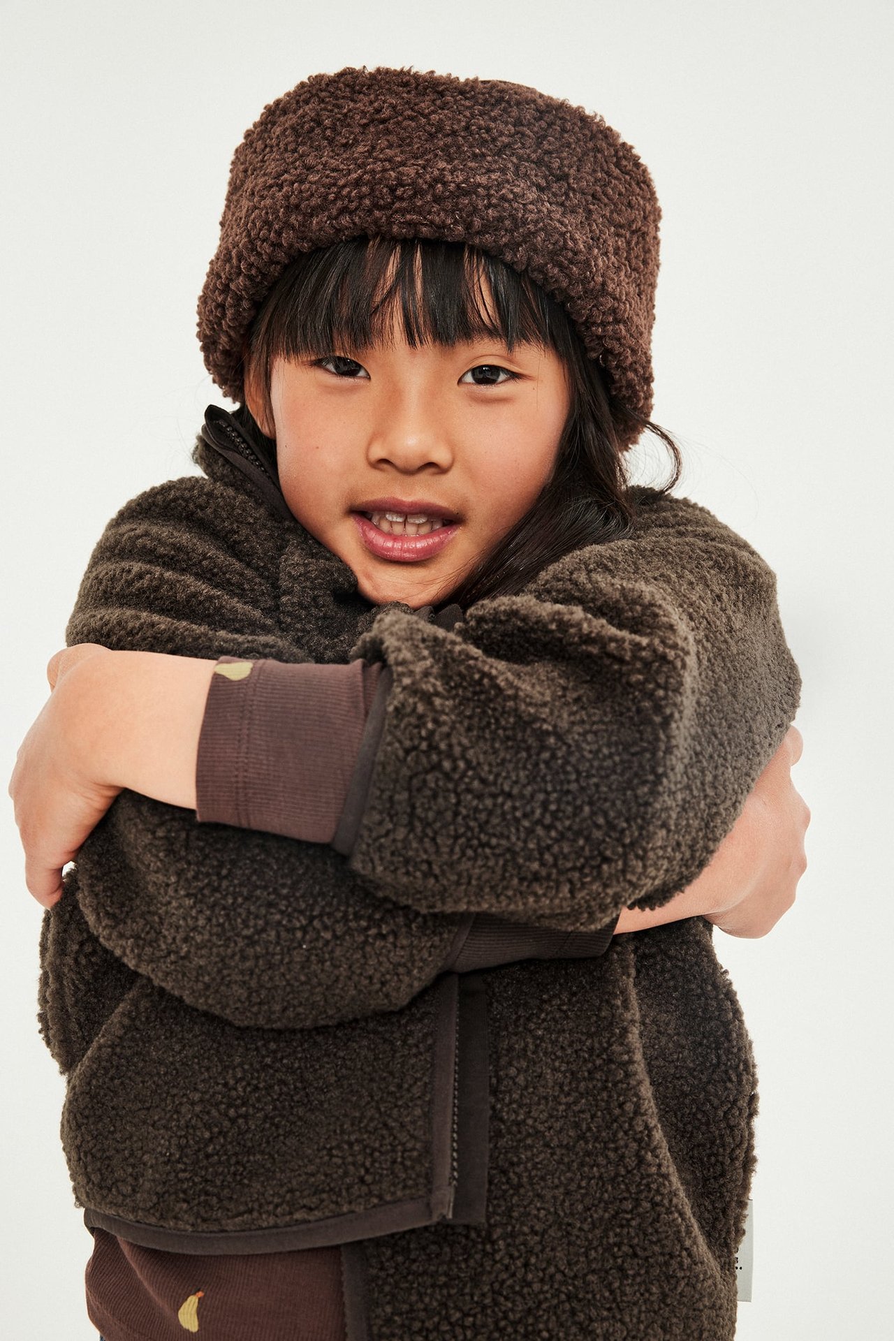 Frontvisning av barn i mørkbrun teddyfleece-jakke, lodden lue og langermet genser.