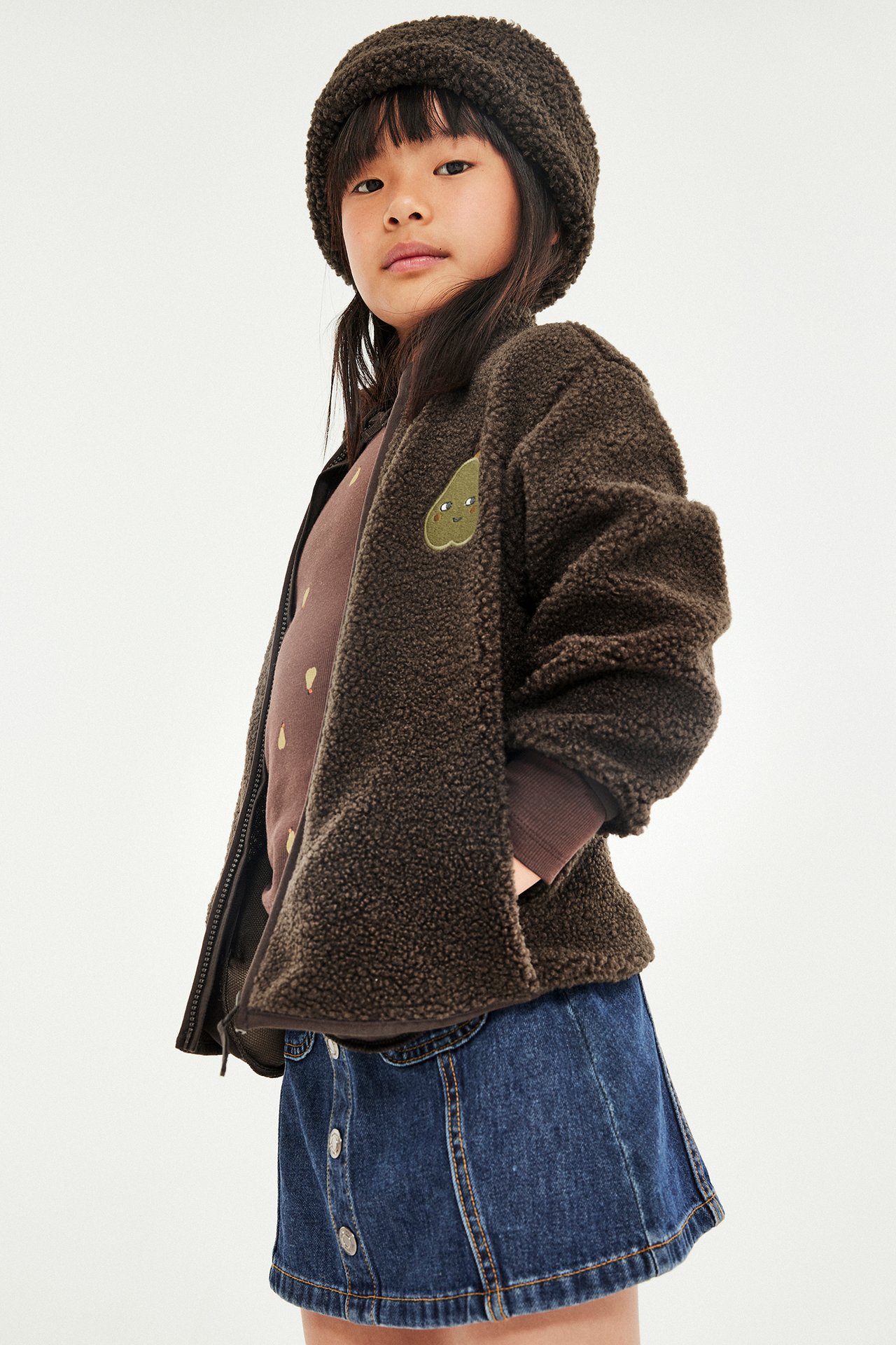 Forfra: Barn har på seg brun teddyfleece-jakke med glidelås, brunprikket topp, denimknappeskjørt og brun teddyfleece-lue.