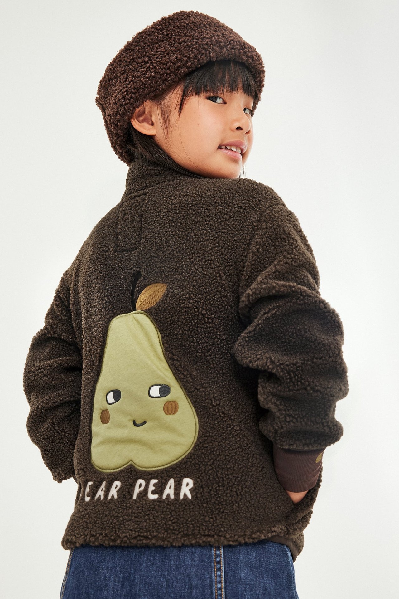 Barn i brun fleecejakke med grønn pæreapplikasjon og 'PEAR PEAR' tekst, brun fleecehatt.