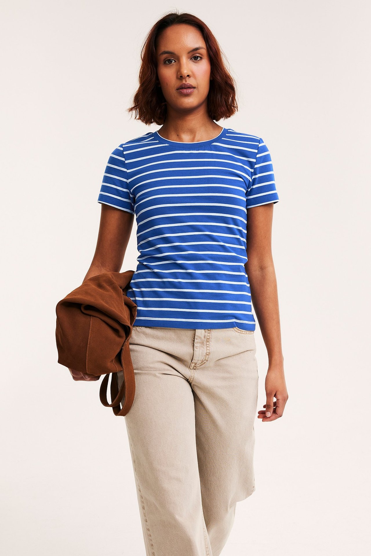 Frontbilde: kvinne i tettsittende blå- og hvitstripet t-skjorte, beige bukse, holder en brun veske.