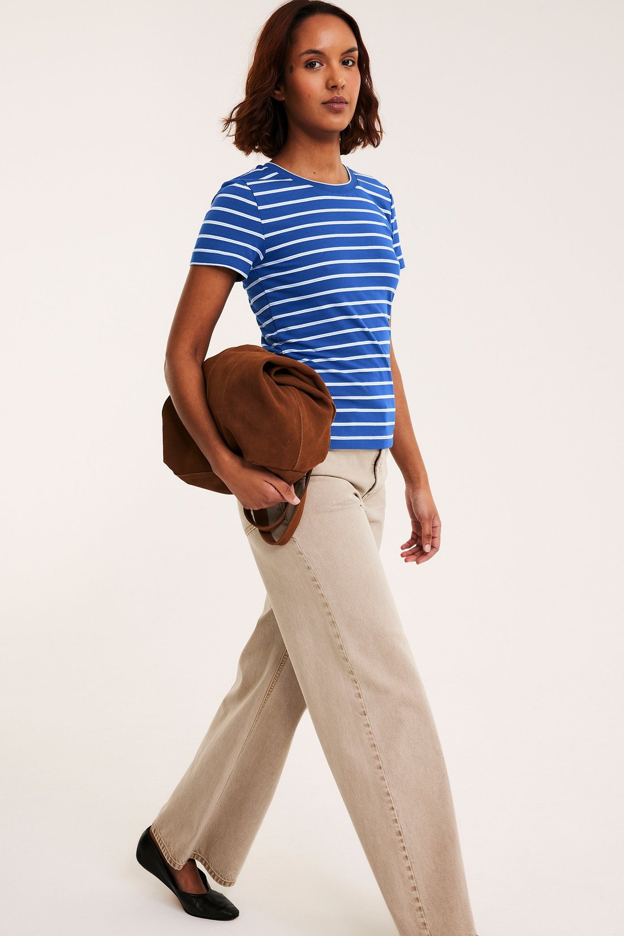 Widok z przodu: Kobieta w dopasowanej niebieskiej koszulce w paski, beżowe szerokie spodnie, czarne baleriny, trzyma brązową torebkę.