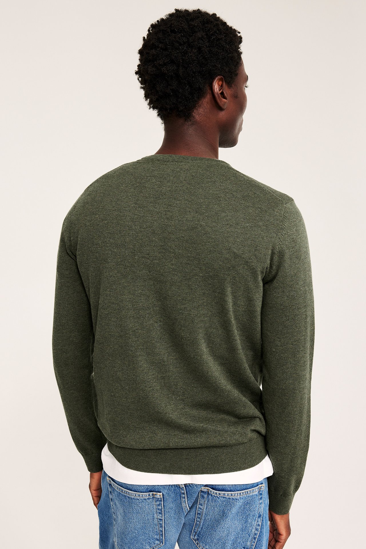 Bakfra av en mann i mørkegrønn genser med rund hals, hvit t-skjorte og blå jeans.
