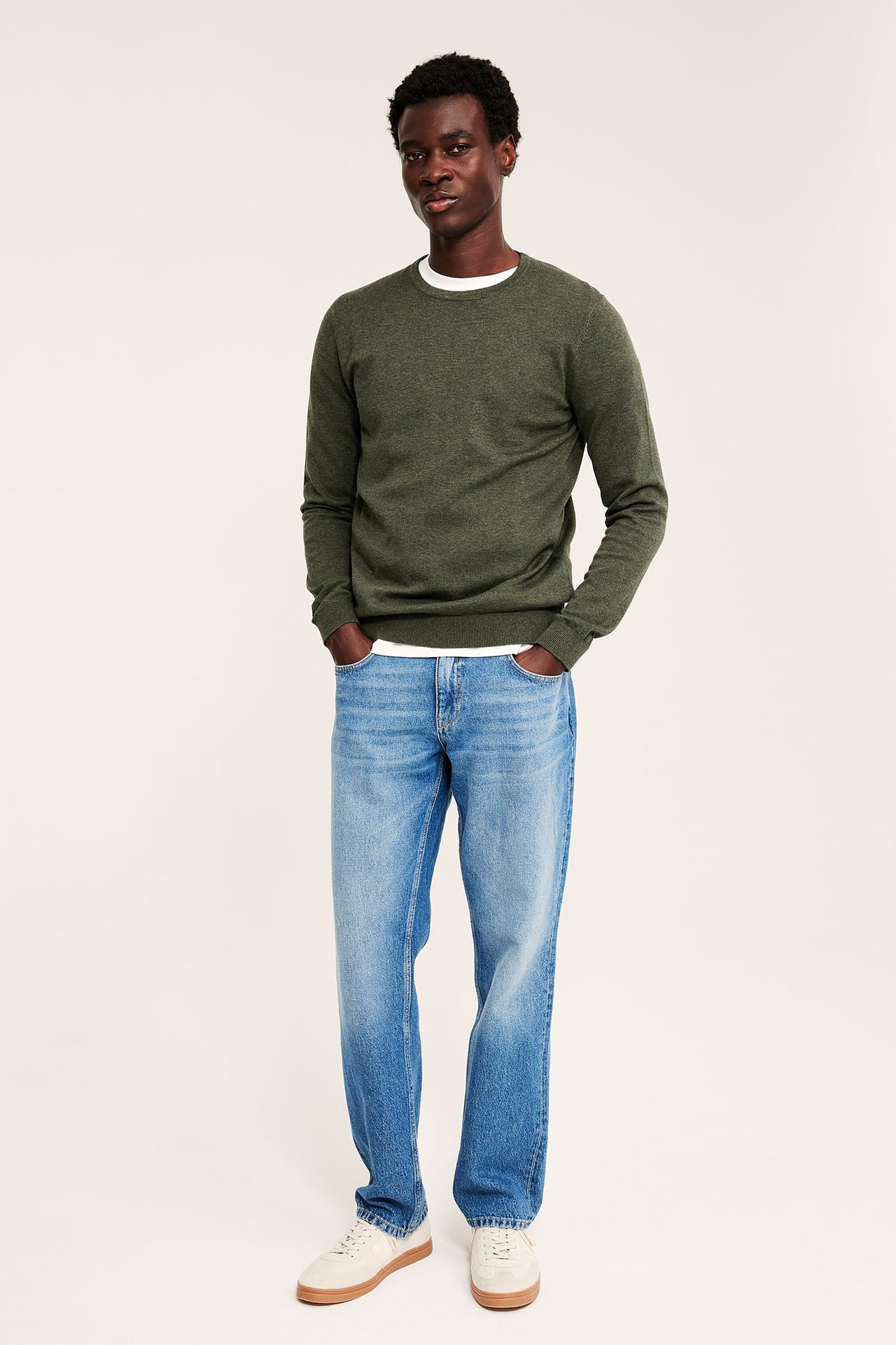 Front: Mannlig modell i grønn crew neck genser, hvit t-skjorte, blå jeans og beige sneakers.