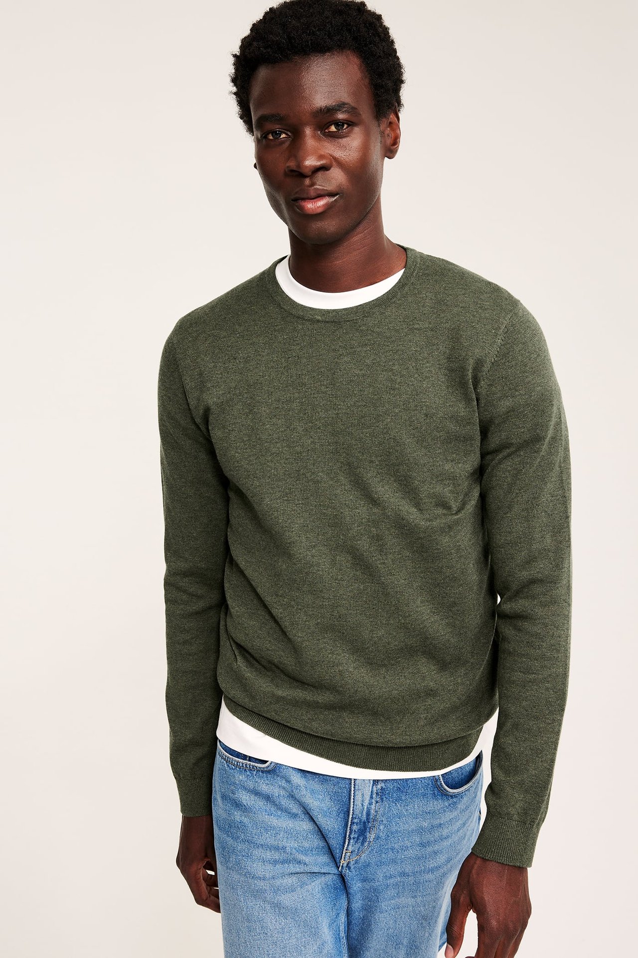 Voksen mann i mørkegrønn genser over hvit t-skjorte og blå jeans, forfra.