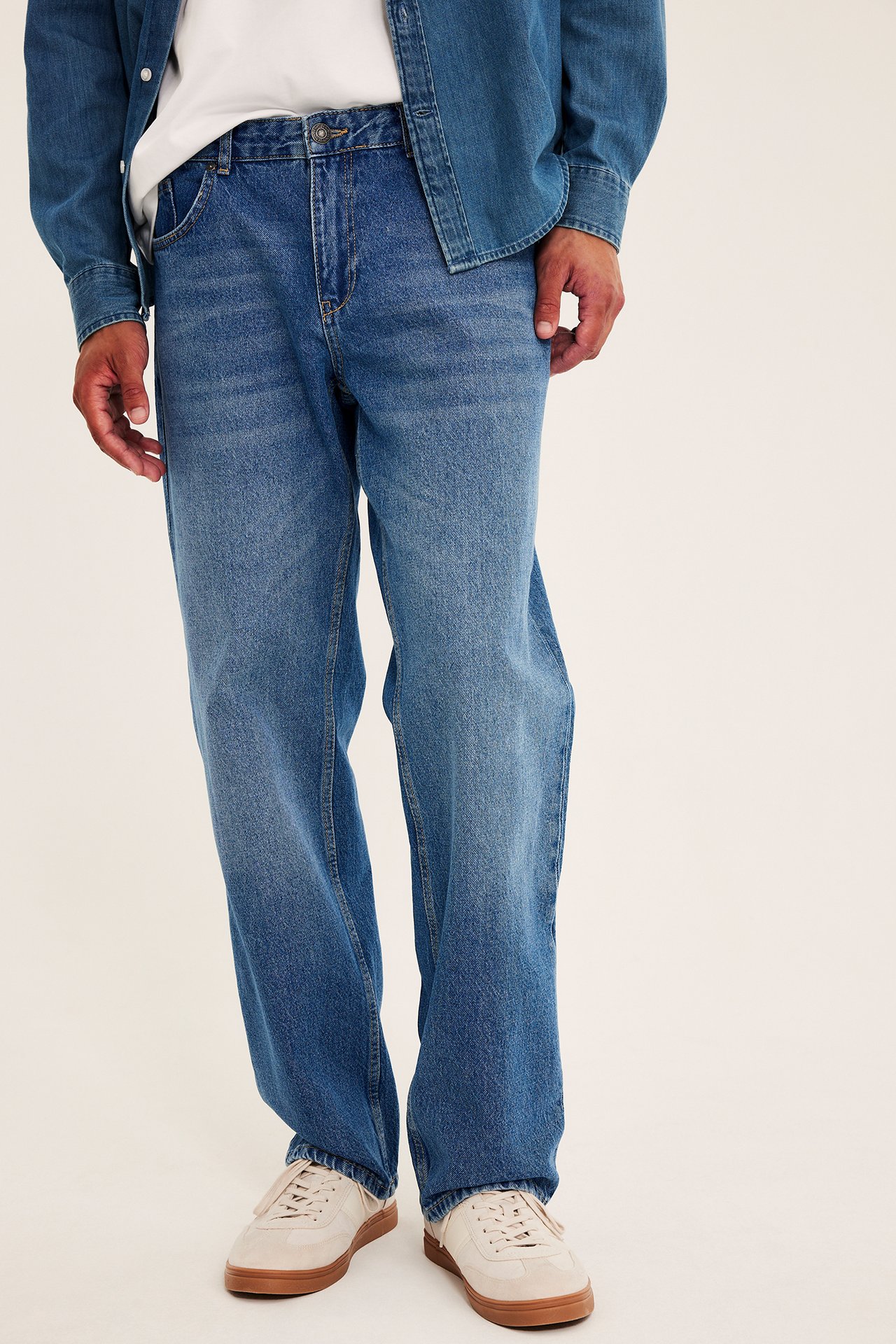 Frontalt bilde av løse blå denimjeans for menn, stylet med en blå denimskjorte og beige joggesko.