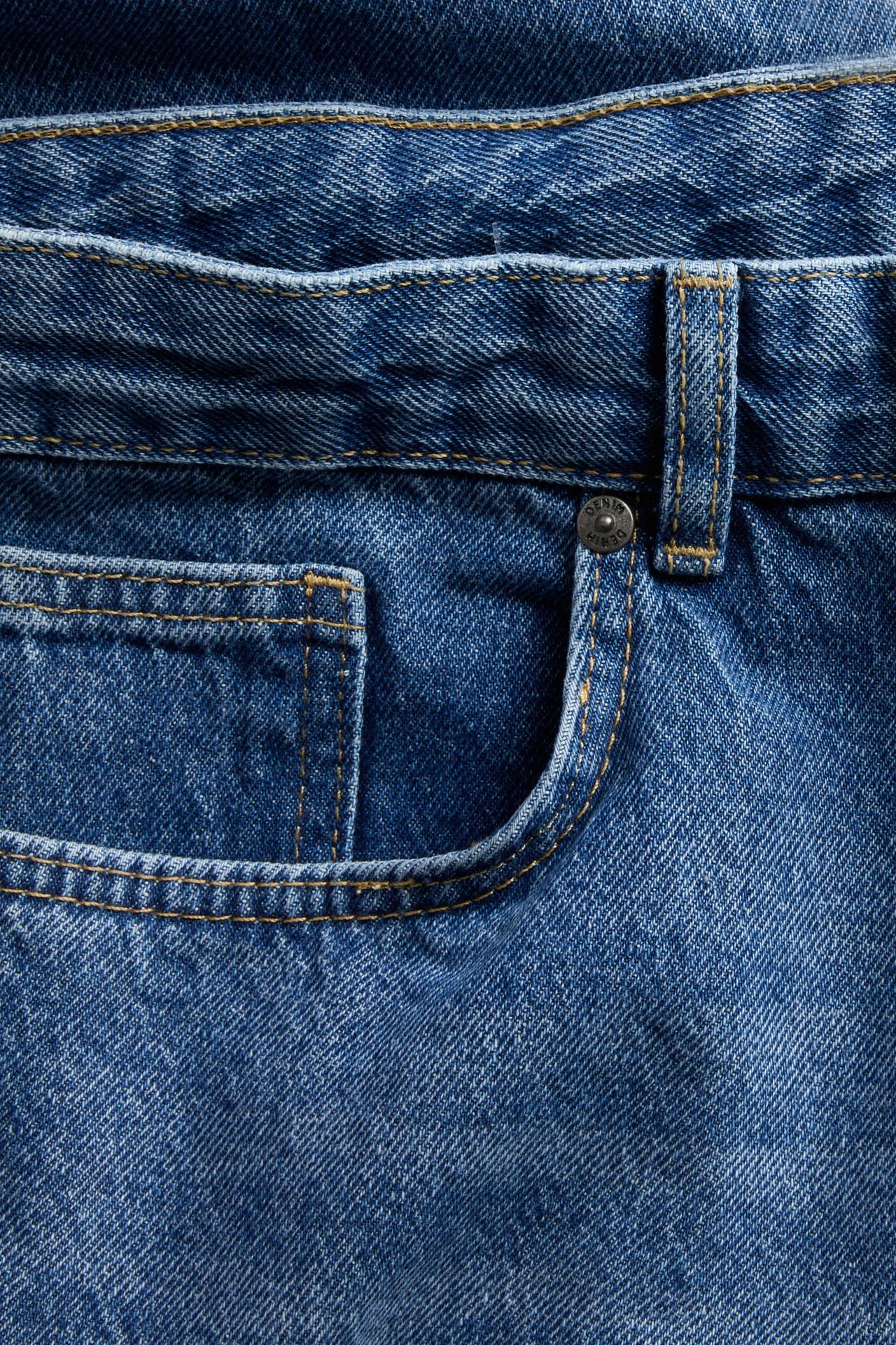 Nærbilde av blå denimjeans med gule sømmer og en metallknapp.