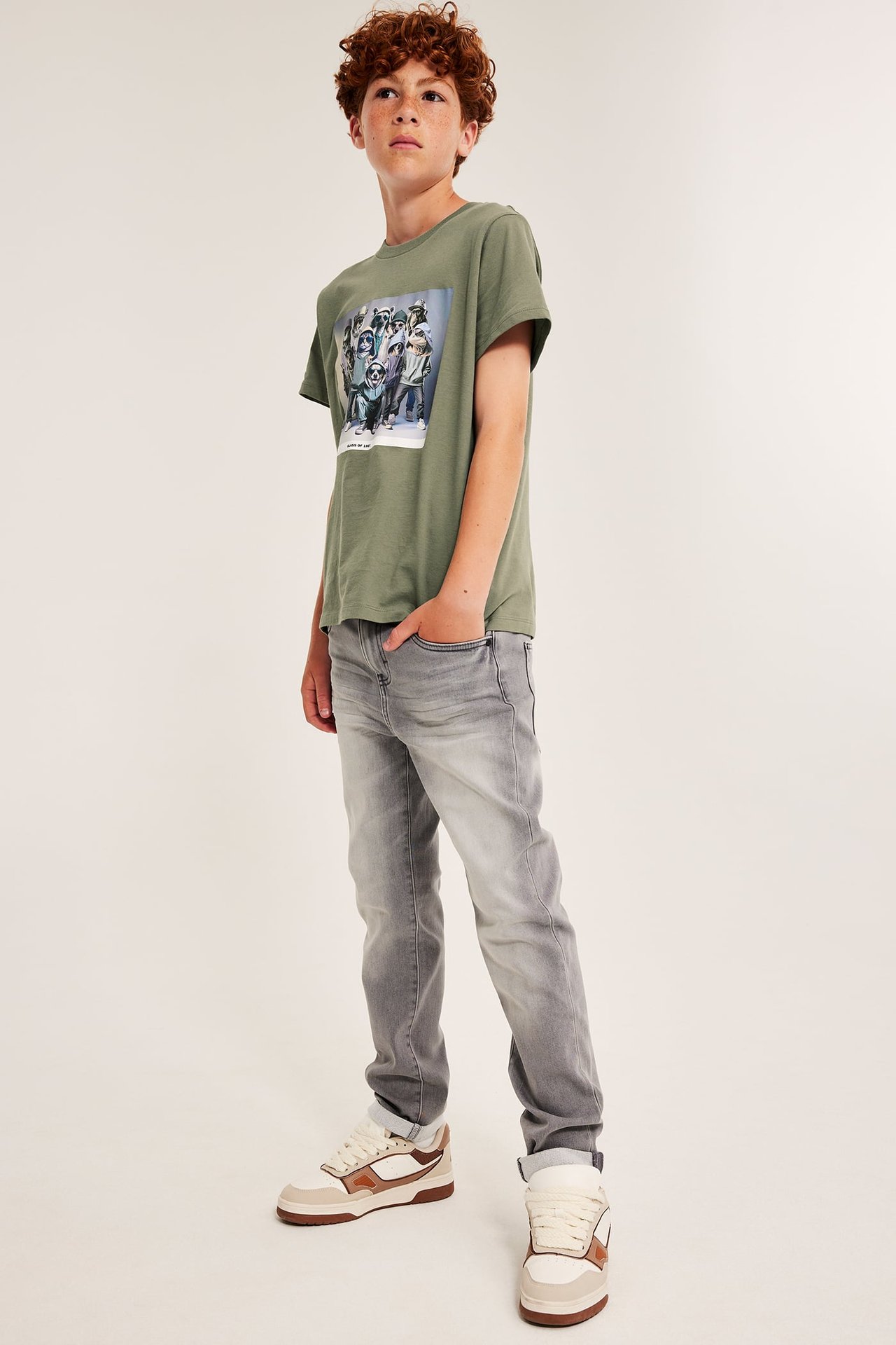 Framifrån, grön t-shirt med grafiskt tryck för barn, kombinerad med grå jeans och beiga och bruna sneakers.