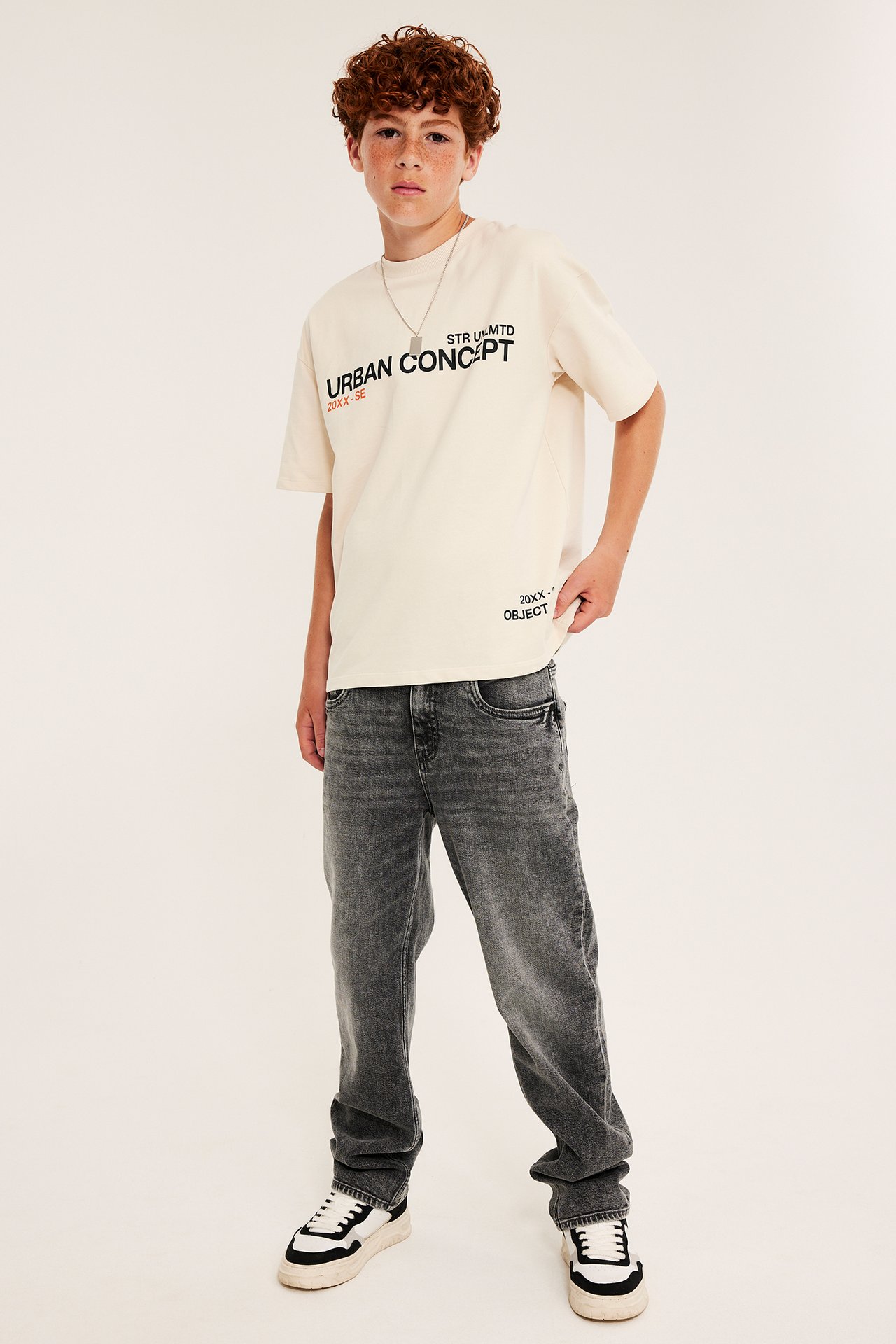En pojke bär en krämfärgad tryckt t-shirt, mörkgrå jeans, svart/vita sneakers och silverhalsband.