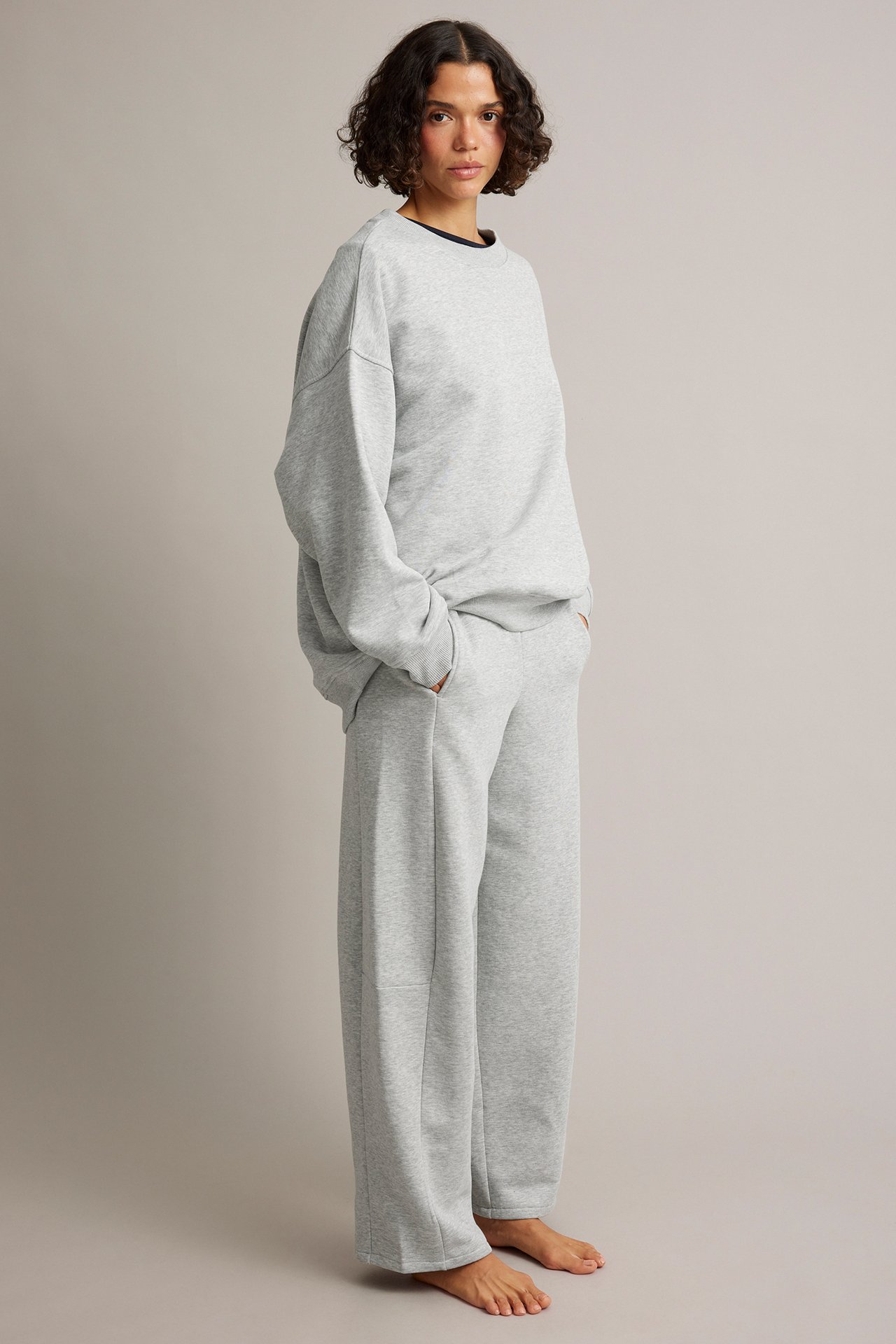 Kvinnlig modell i ljusgrå oversize sweatshirt och vida sweatpants, med händerna i fickorna.