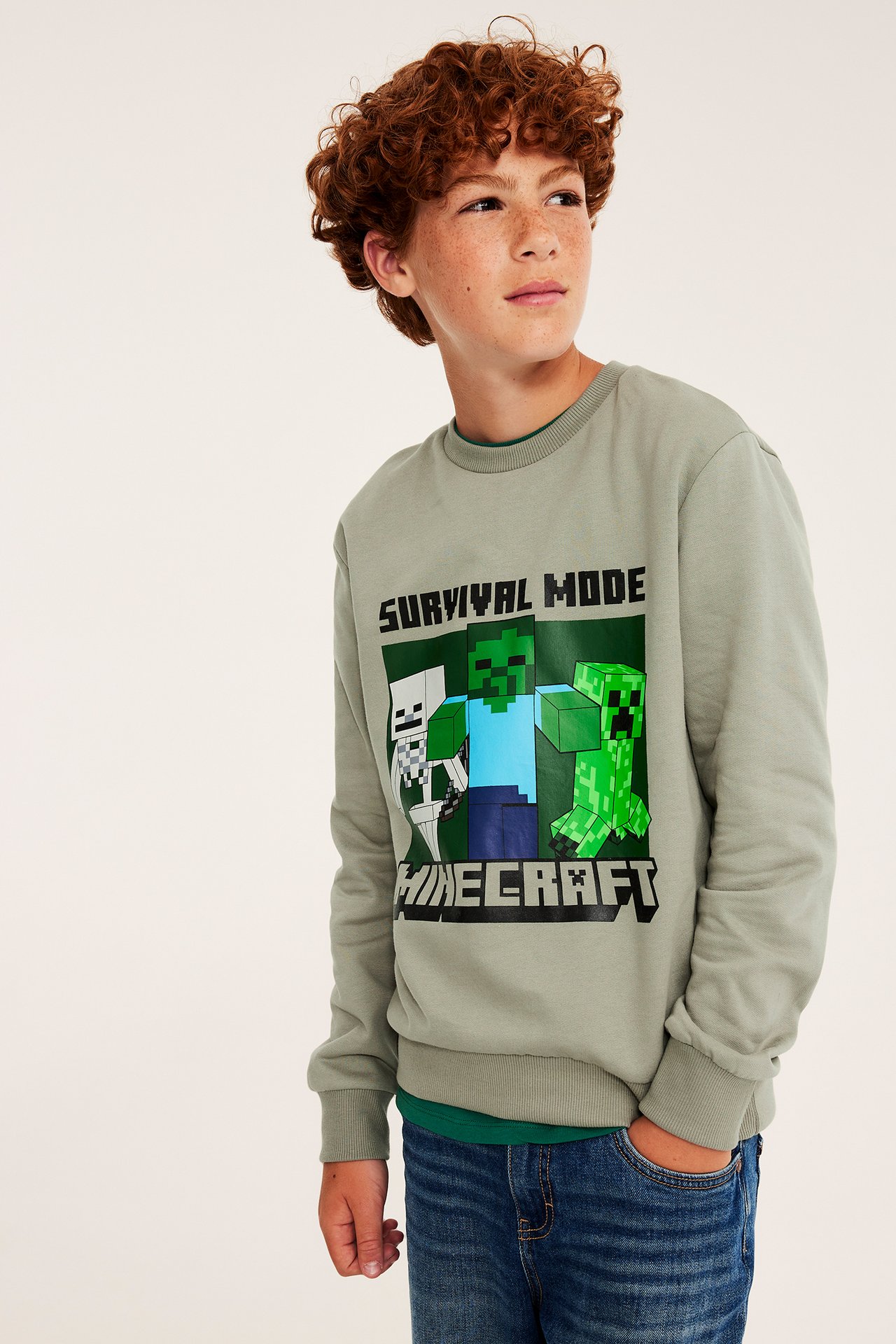 Edestä näkymä lapsesta salvianvihreässä 'Survival Mode Minecraft' collegepaidassa ja sinisissä farkuissa.