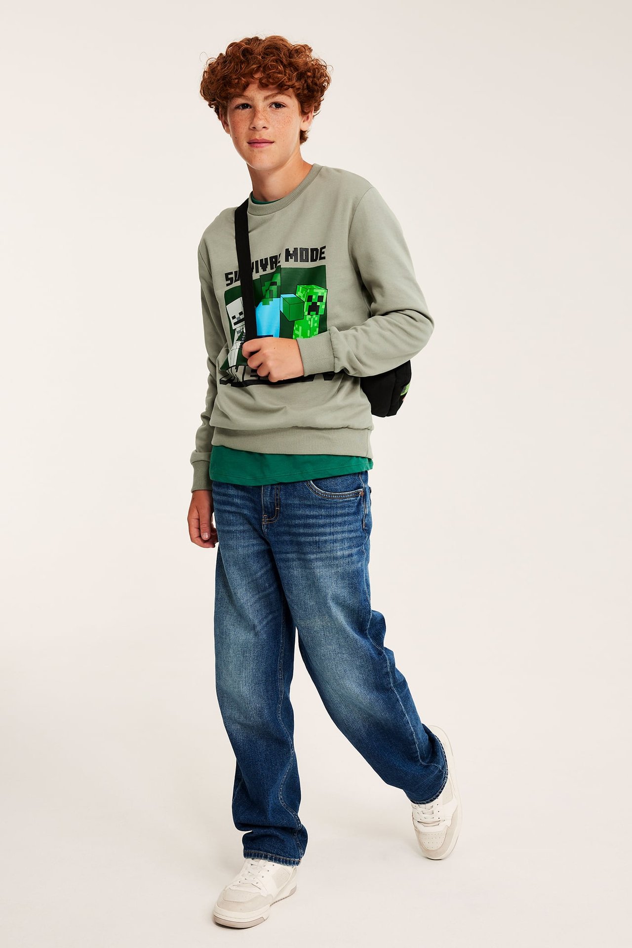 Widok z przodu, chłopiec w szarozielonej bluzie Minecraft, zielonym t-shircie, niebieskich dżinsach, białych tenisówkach i czarnej torbie.