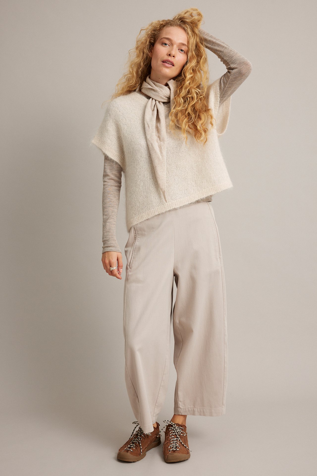 Frontbilde av kvinne i lys beige strikket poncho, taupe langermet topp, cropped bukser og brune sko.