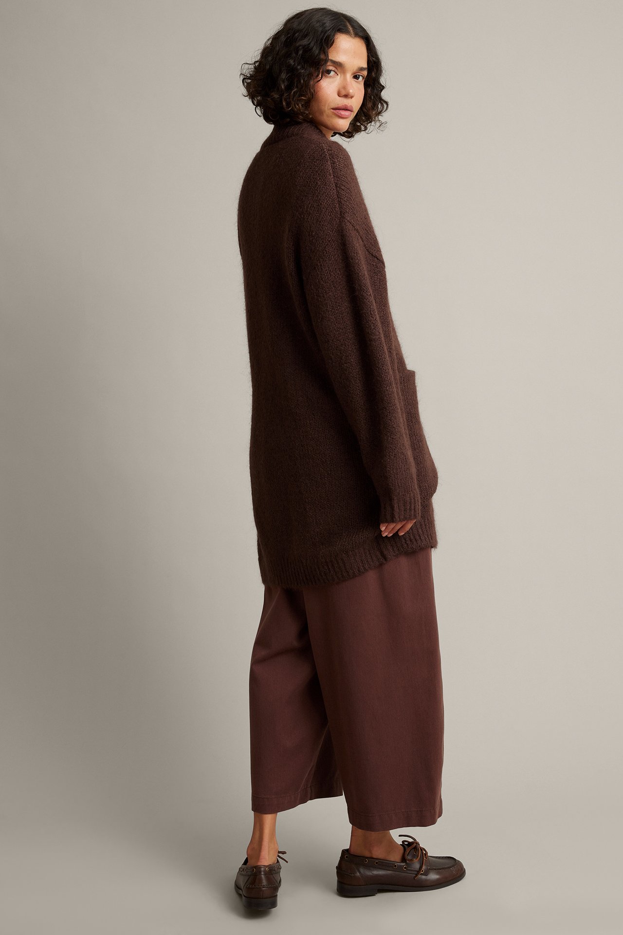 Sidevisning, kvinnelig modell i brun strikket cardigan, brune vide bukser og mørkebrune loafers.