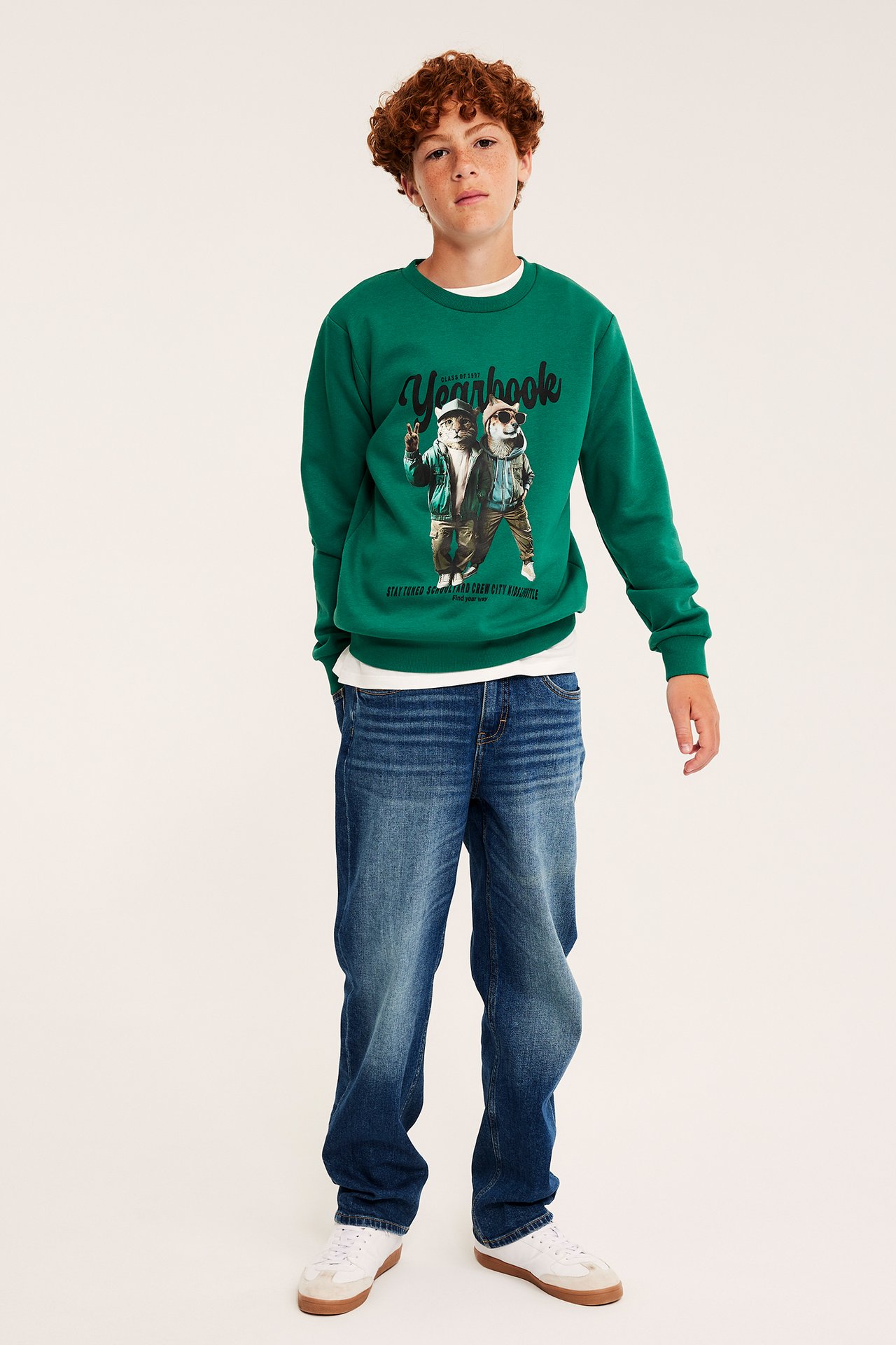Pojke framifrån i grön grafisk sweatshirt, blå jeans och vita sneakers.