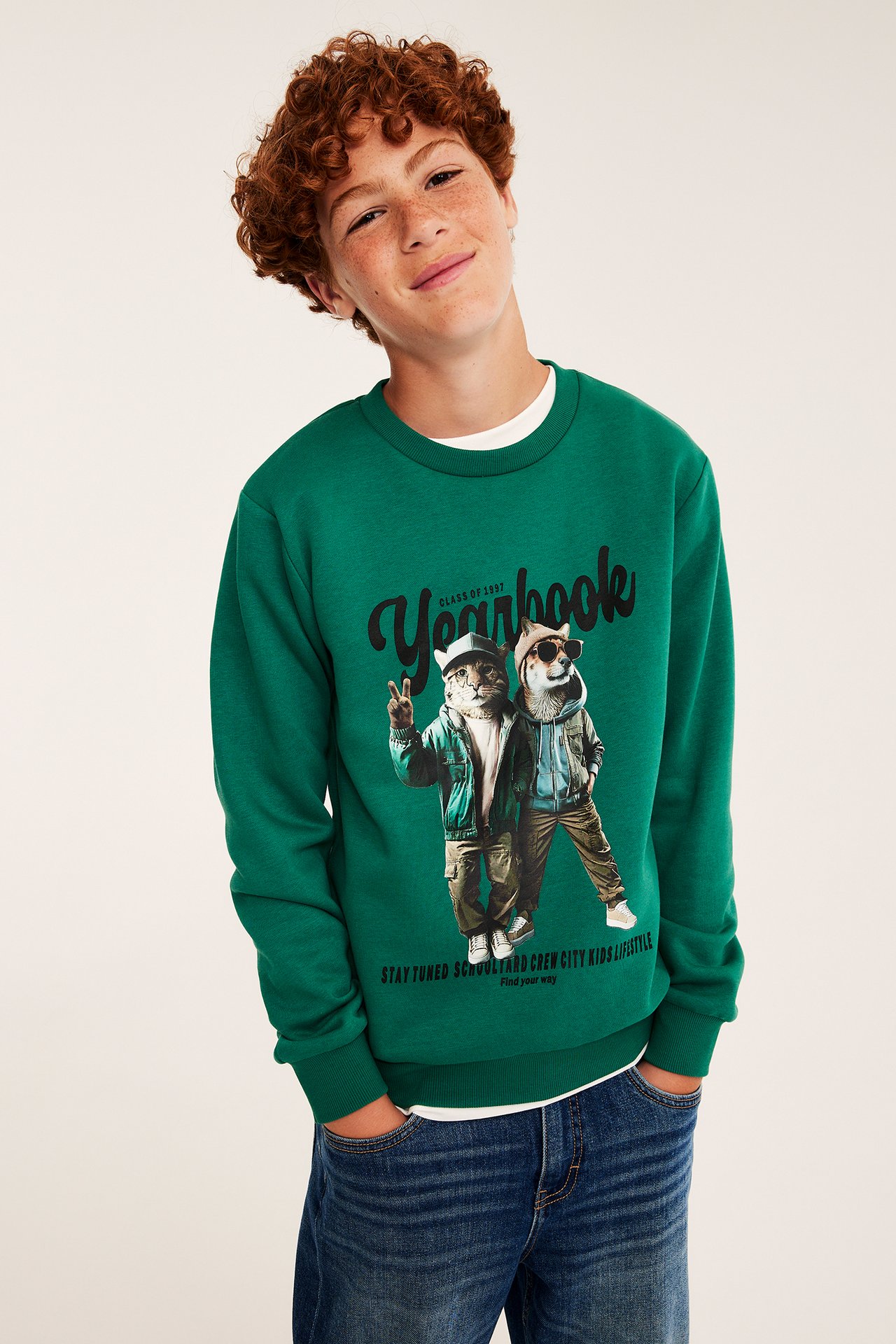 Frontvy av ett barn i en grön crewneck-tröja med djurmönster och blåa jeans.