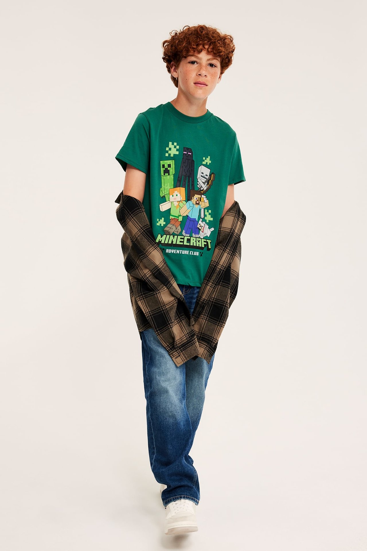 Framsida av ett barn i grön Minecraft t-shirt, brunrutig skjorta, blå jeans och vita sneakers.
