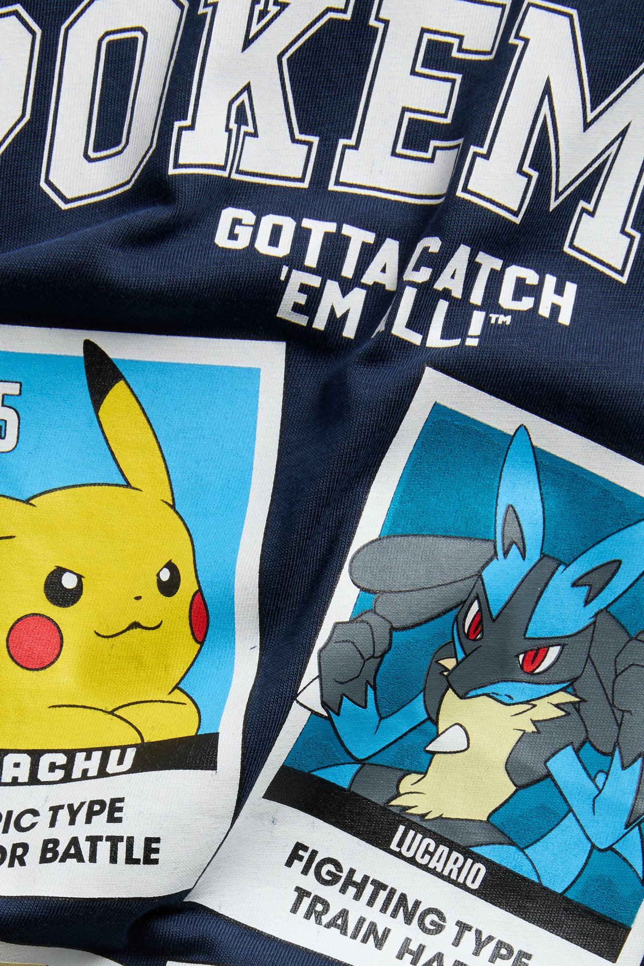 Lähikuva: Tummasininen t-paita, jossa valkoinen POKEMON-logo, 'Gotta Catch 'Em All!', Pikachu- ja Lucario-kuvat.