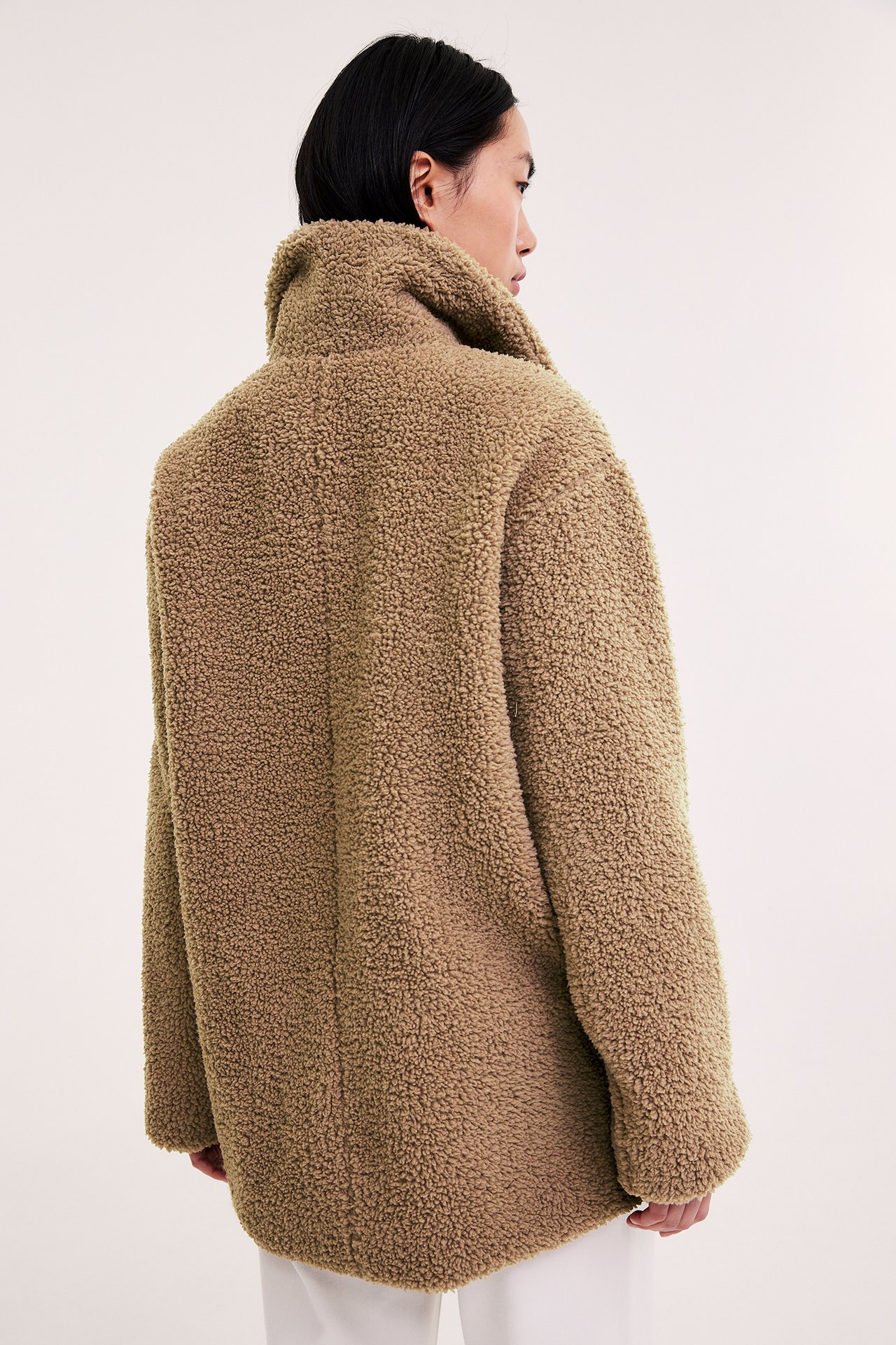 Baksidebilde av en kvinne i en løstsittende, mellomlang beige pilejakke over hvite bukser.