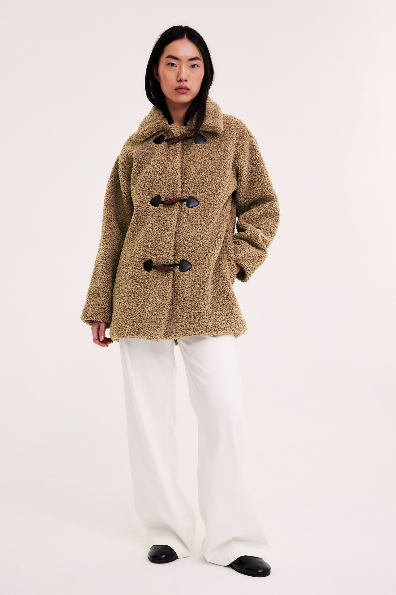 Etunäkymä: Nainen beigessä puolipitkässä nallepuserossa, jossa duffelikiinnitys, valkoiset housut ja mustat kengät.