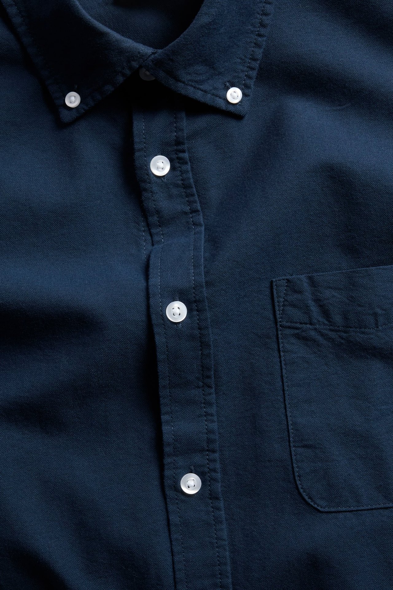 Nærbilde av en mørkeblå skjorte med button down-krage, hvite knapper og brystlomme.