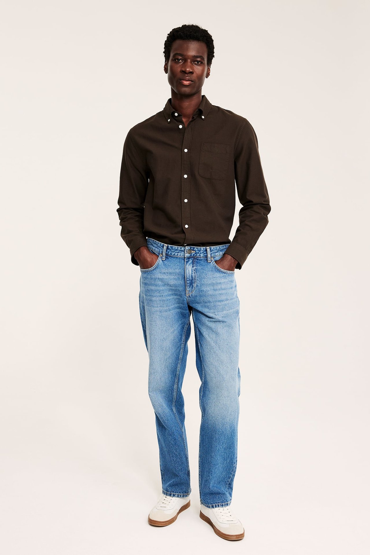 Frontbilde av mann i mørkebrun langermet skjorte, blå jeans og hvite joggesko.