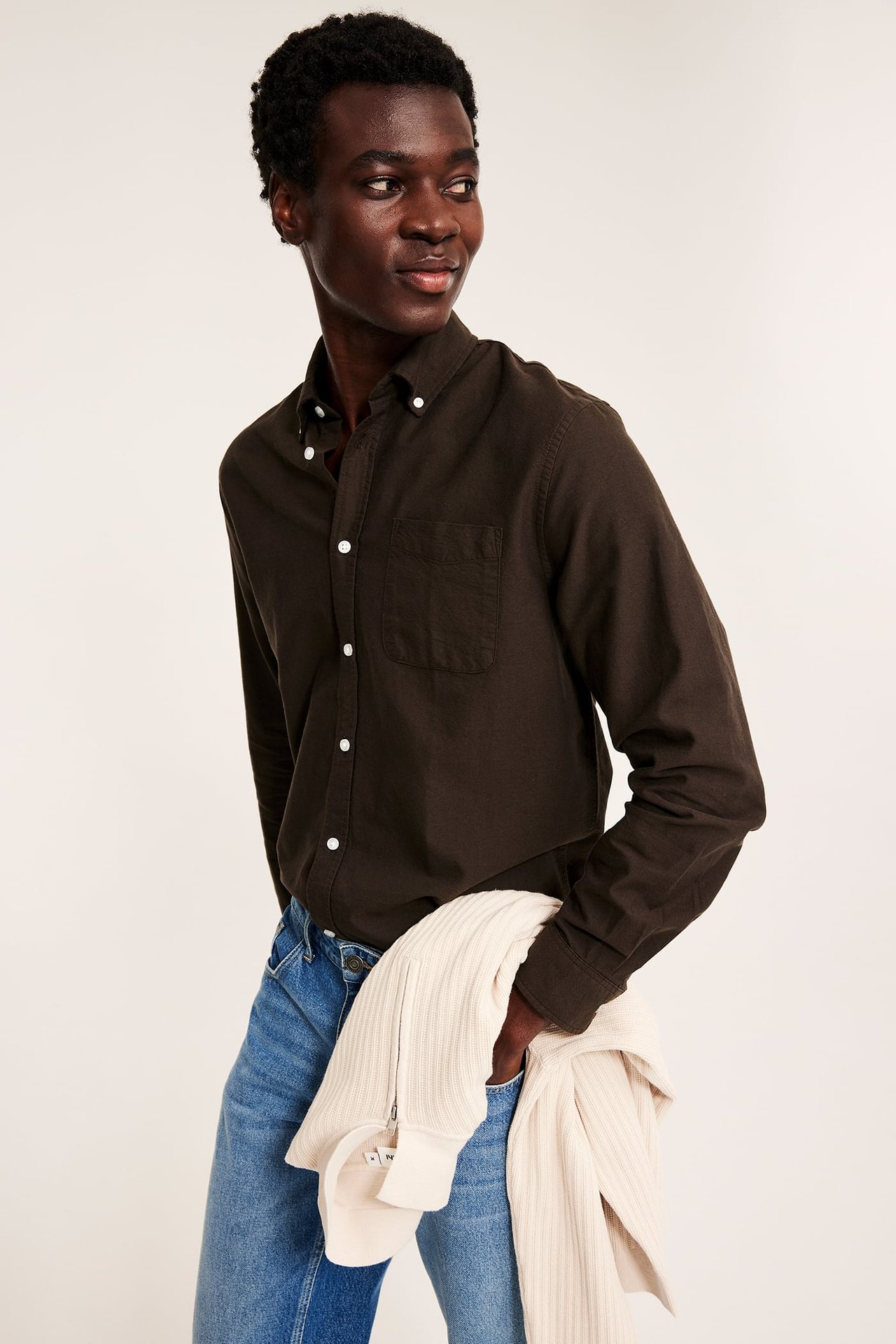 Mannlig modell har mørkebrun skjorte, blå jeans, holder lysebeige ribbet glidelåsgenser.