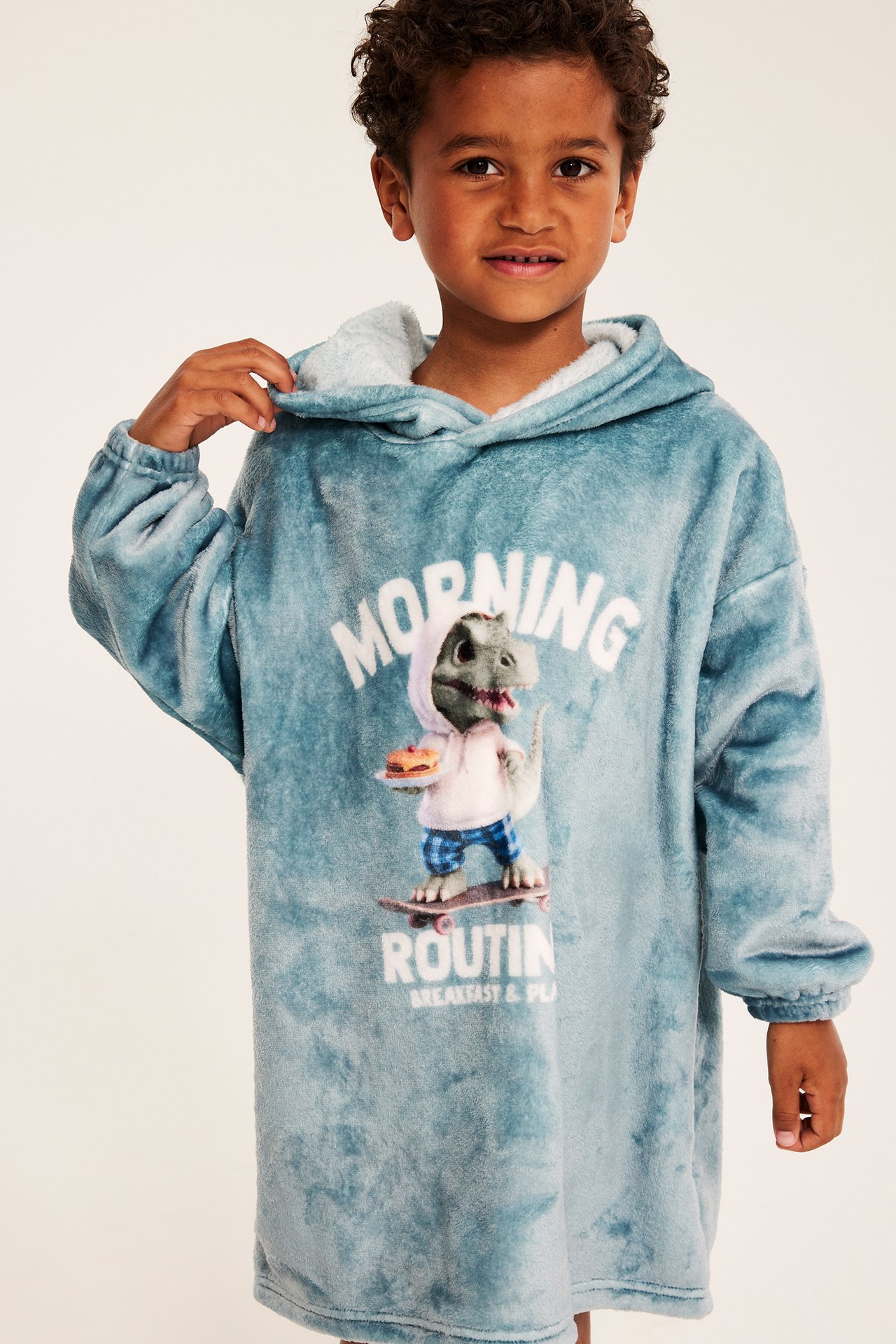 Framifrån, barn i oversized blå fluffig huvtröja med dinosauriemotiv.