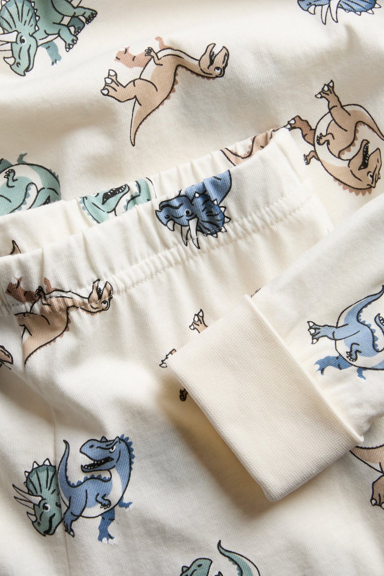 Lähikuva lasten luonnonvalkoisesta puuvillapyjamasta dinosauruskuviolla, joustavalla vyötäröllä ja taitettavilla hihansuilla.