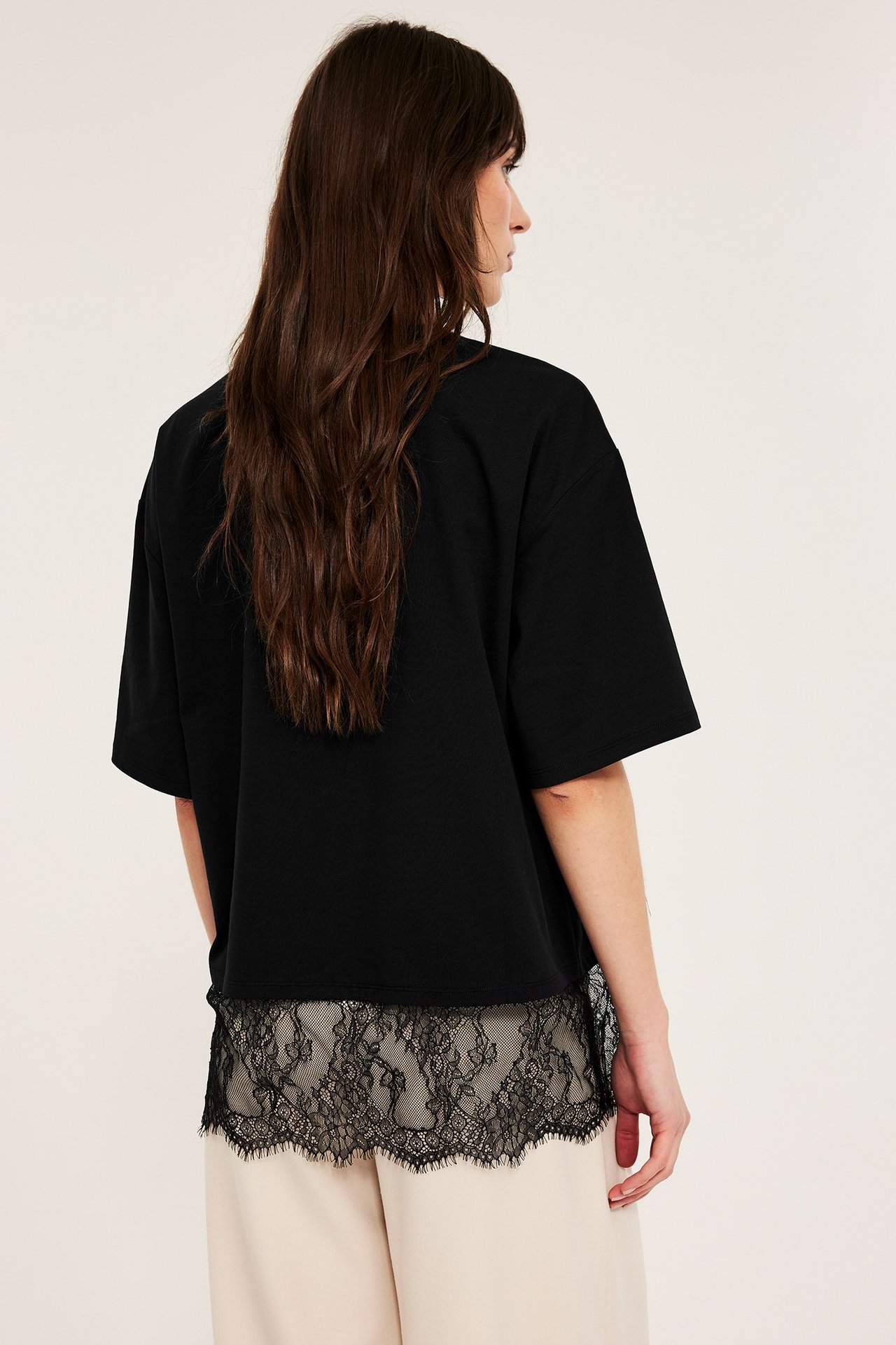 Bakfra av en kvinne i svart løs t-skjorte med dekorativ blondekant over lyse beige bukser.