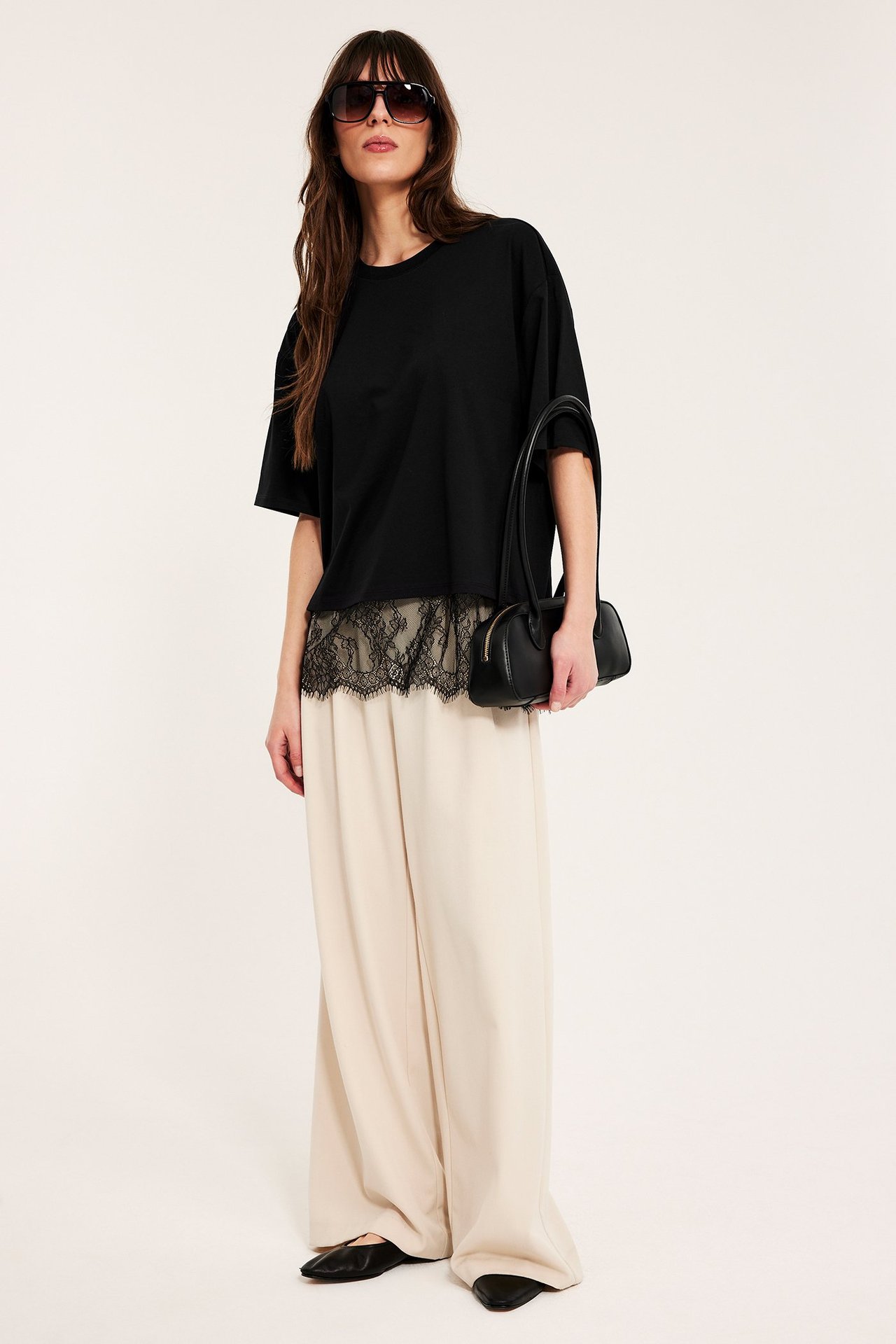 Framifrån: Kvinna i svart lös t-shirt med spetskant, beiga vida byxor, svarta flats, solglasögon, med svart handväska.
