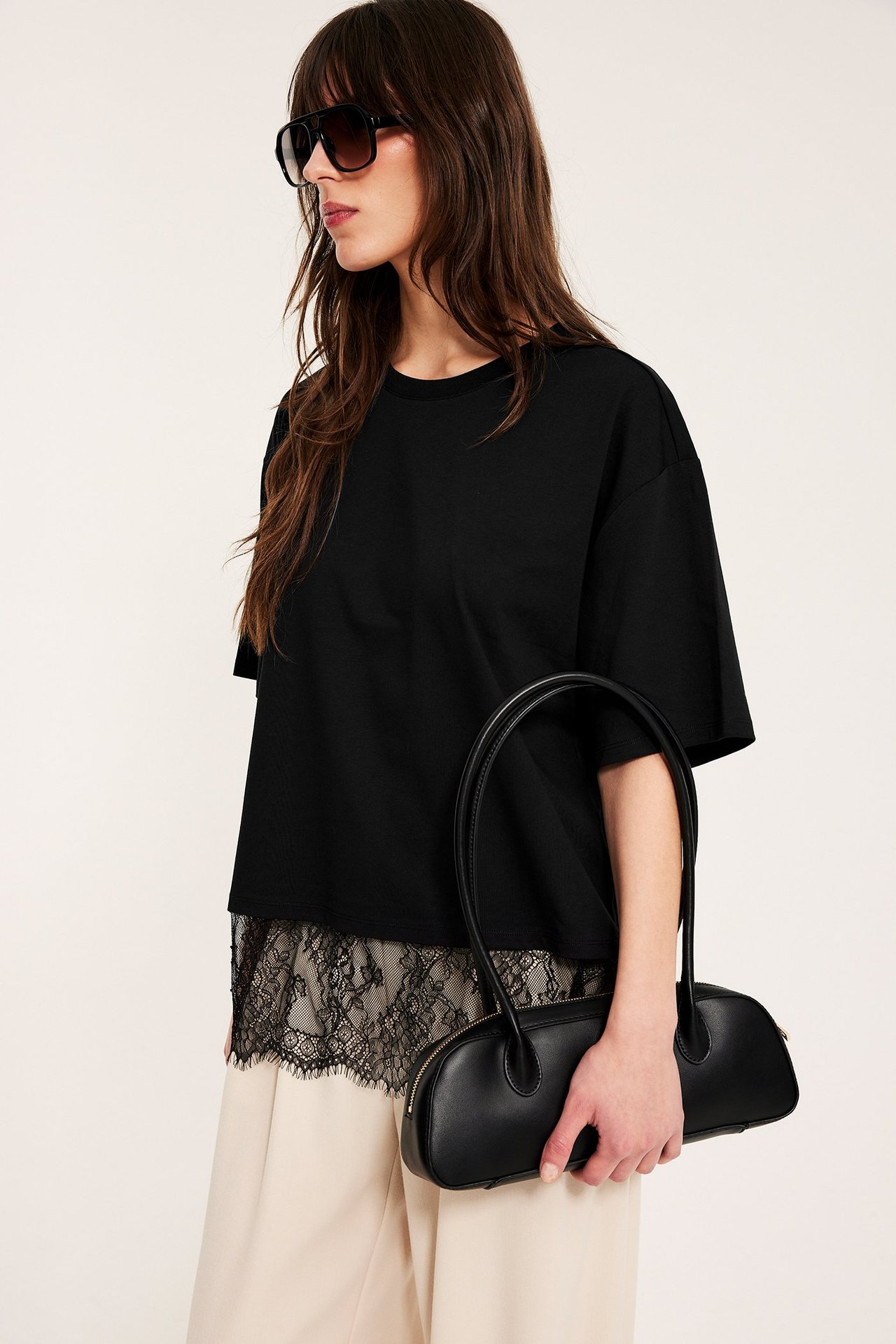 Sidebilde kvinne iført svart løs t-skjorte med blondedetalj, beige bukse, holder svart veske, solbriller.