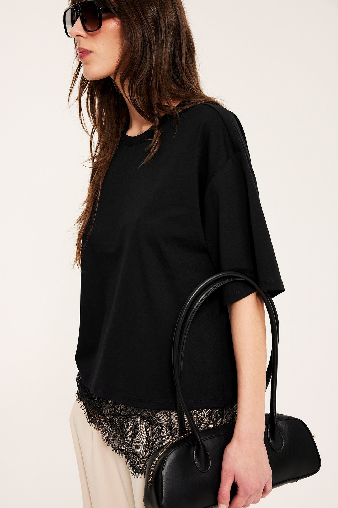 Widok z boku kobiety: luźny czarny t-shirt z koronkowym dołem, jasne spodnie, czarne okulary, czarna torebka.
