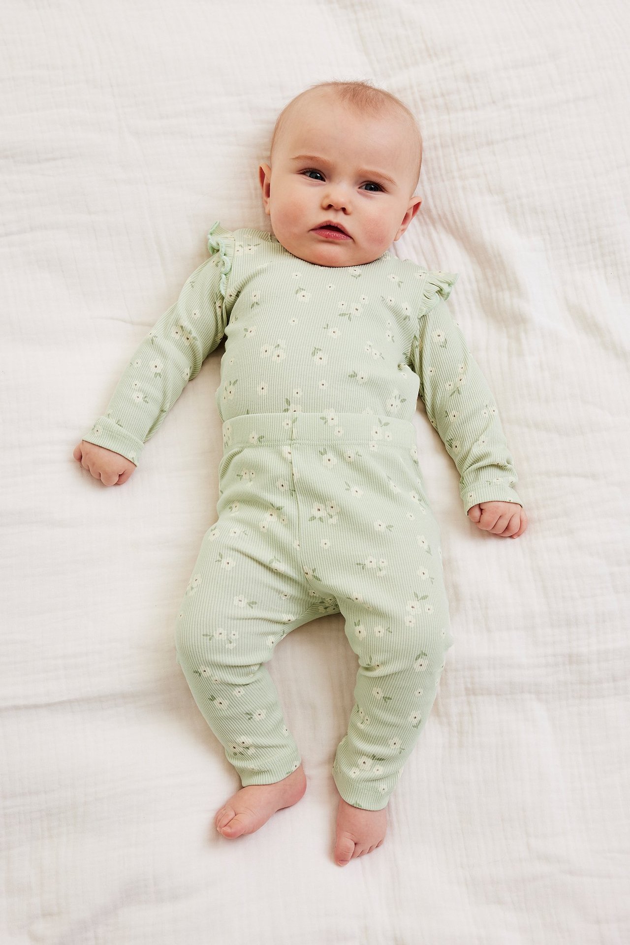 Ylhäältä kuvattu vauva yllään vaaleanvihreä ribbineuleinen kukkakuvioinen puuvillasetti, jossa röyhelöolkapäät.