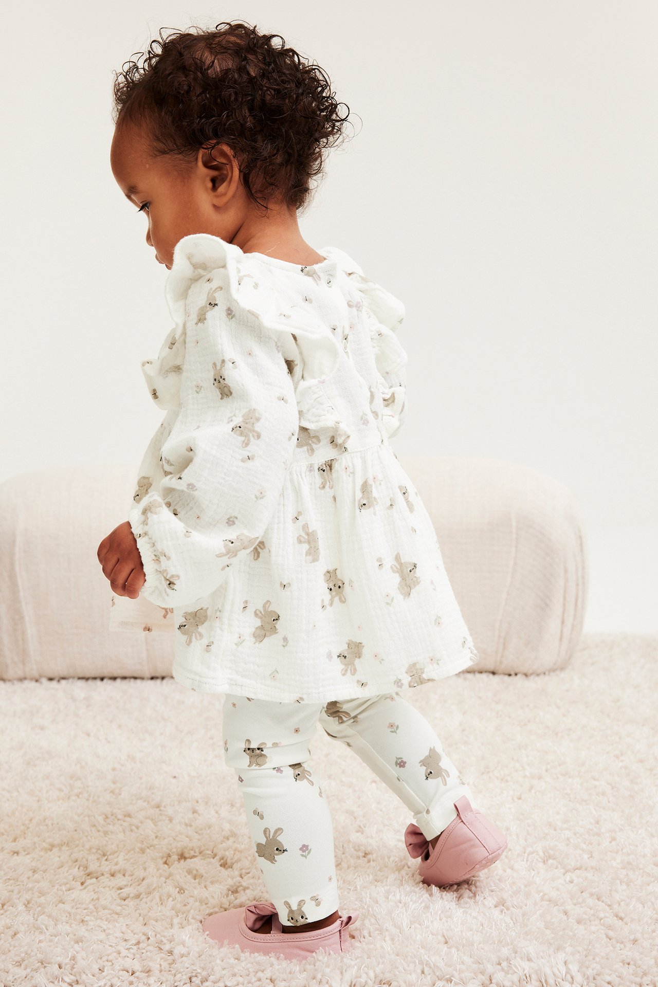 Sidevisning av en baby i offwhite leggings med kaninmønster, matchende rysjet tunika og rosa ballettsko.