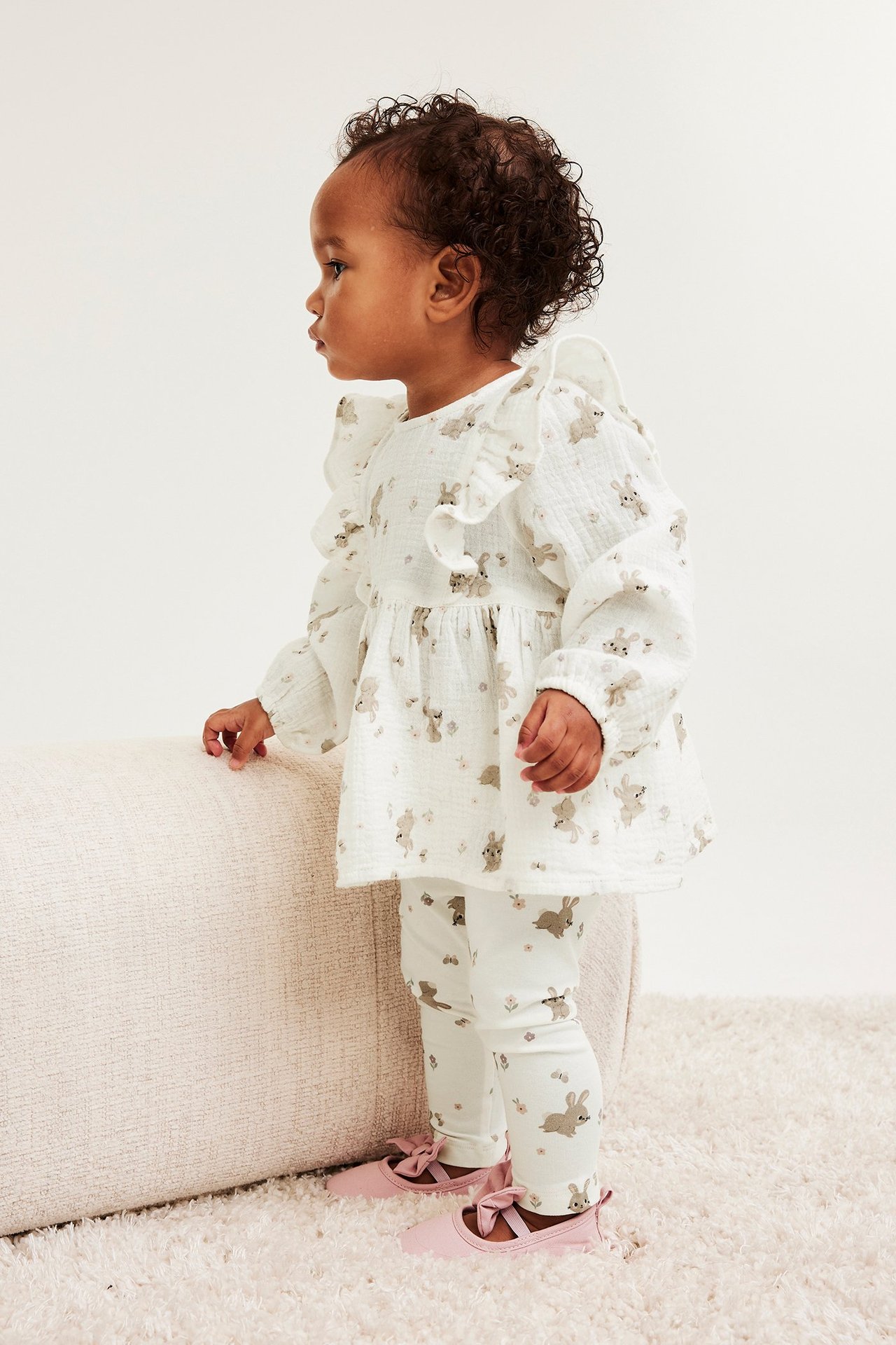 Sivukuva vauvasta, jolla on luonnonvalkoiset pupukuvioiset leggingsit, yhteensopiva röyhelötoppi ja vaaleanpunaiset balettikengät.