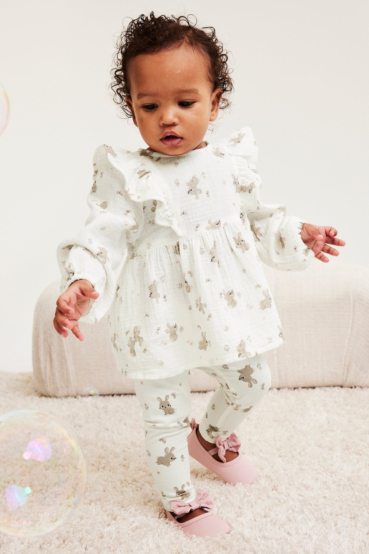 Dziecko z przodu: ecru legginsy w króliczki, pasująca tunika z falbankami i różowe buciki z kokardkami.