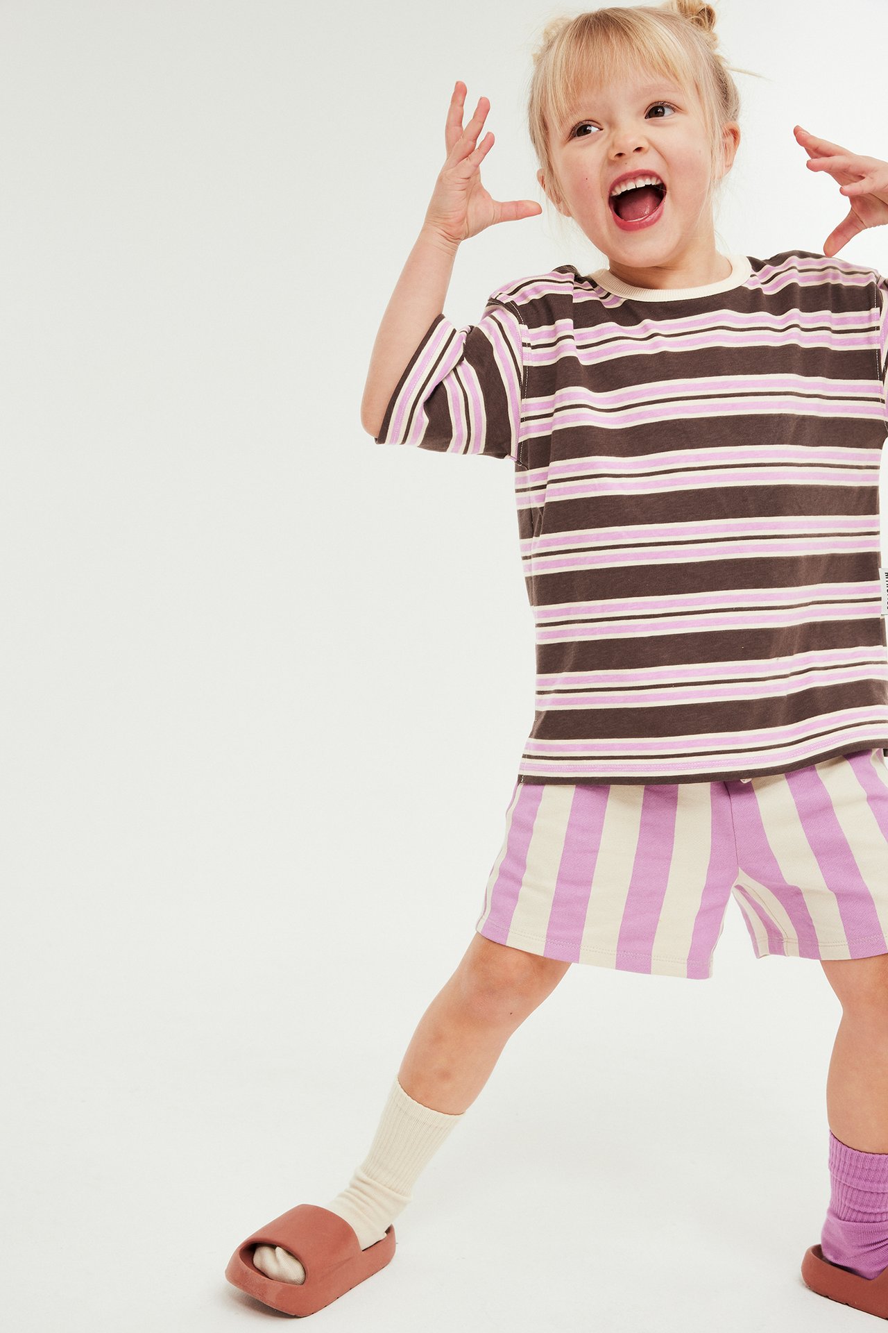 Barn har på seg brun, rosa, beige stripet t-skjorte, lilla stripete shorts, tofargede sokker og brune sandaler.