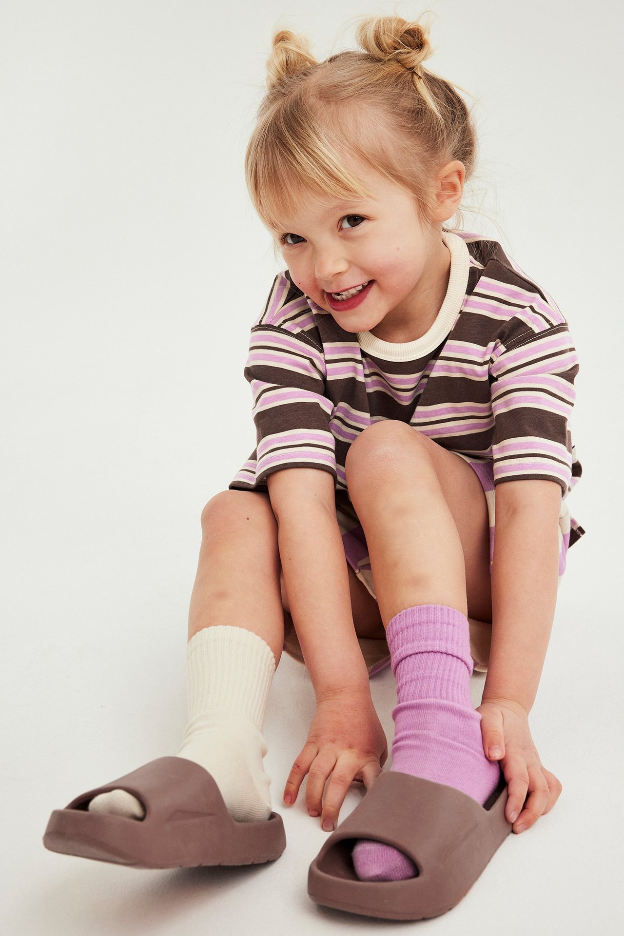 Tyttö pukeutuu ruskea-vaaleanpunaiseen raidalliseen t-paitaan, liila shortsit, erivärisiin sukkiin ja ruskeisiin sandaaleihin.