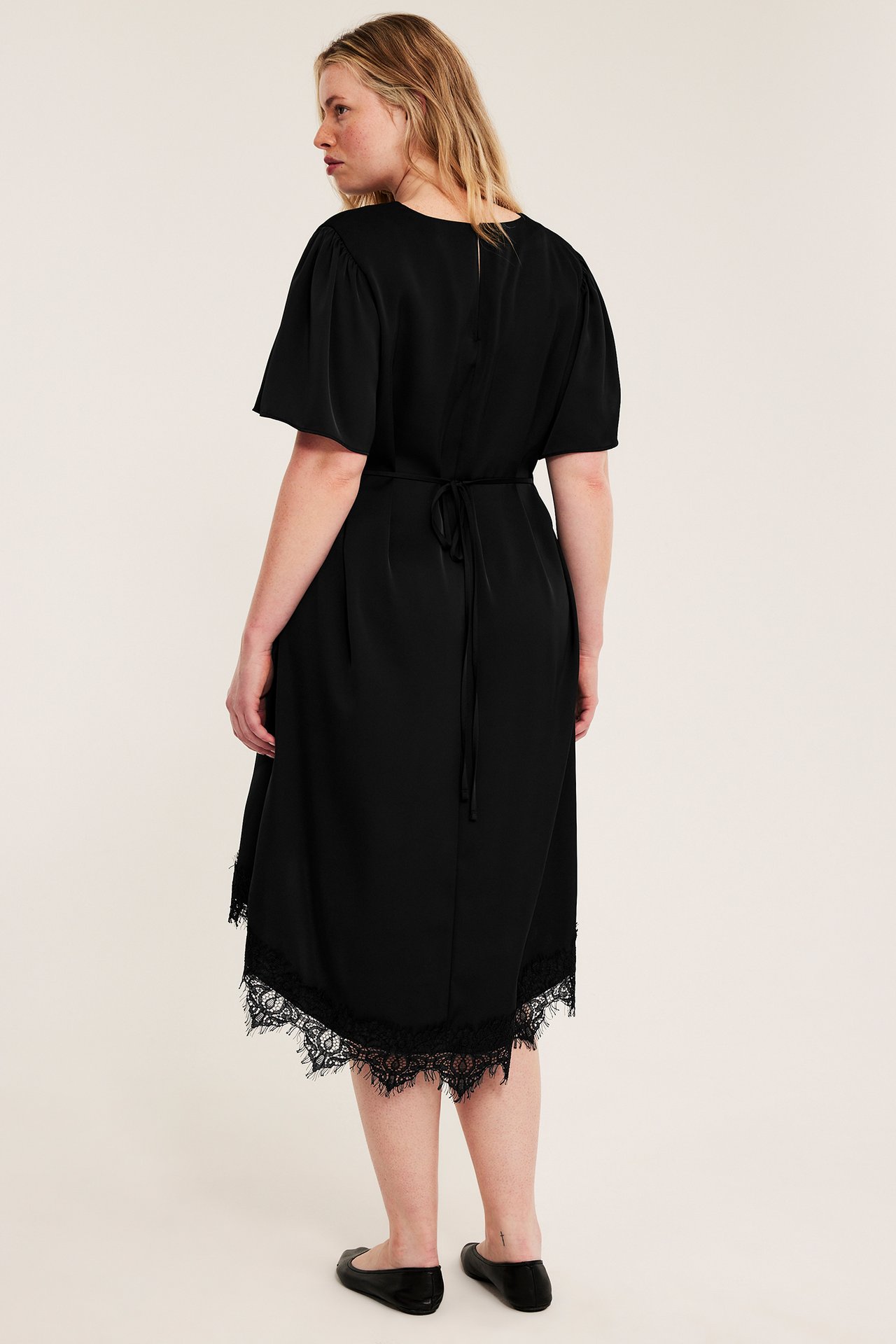 Widok z tyłu: kobieta w czarnej satynowej sukience z krótkimi rękawami, wiązaną talią, koronką i czarnymi baletkami.