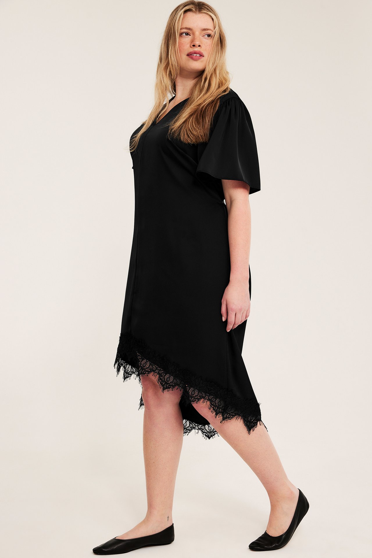 Sidovy: Kvinna i svart satinklänning med korta, vida ärmar, asymmetrisk spetsfåll och svarta ballerinaskor.
