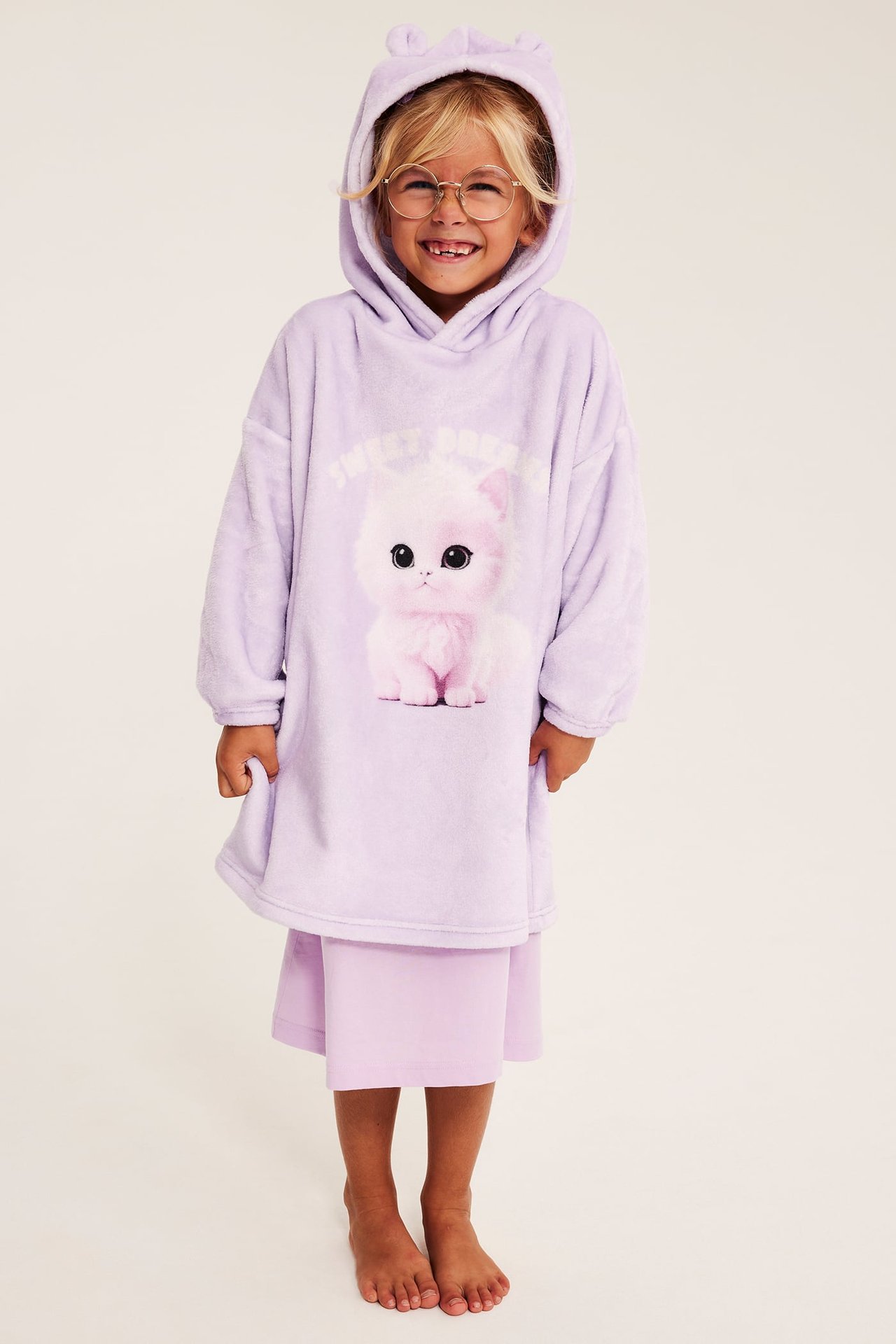 Framifrån: Barn i lila fluffigt nattlinne med huva, kattöron och tryck, över lila shorts.