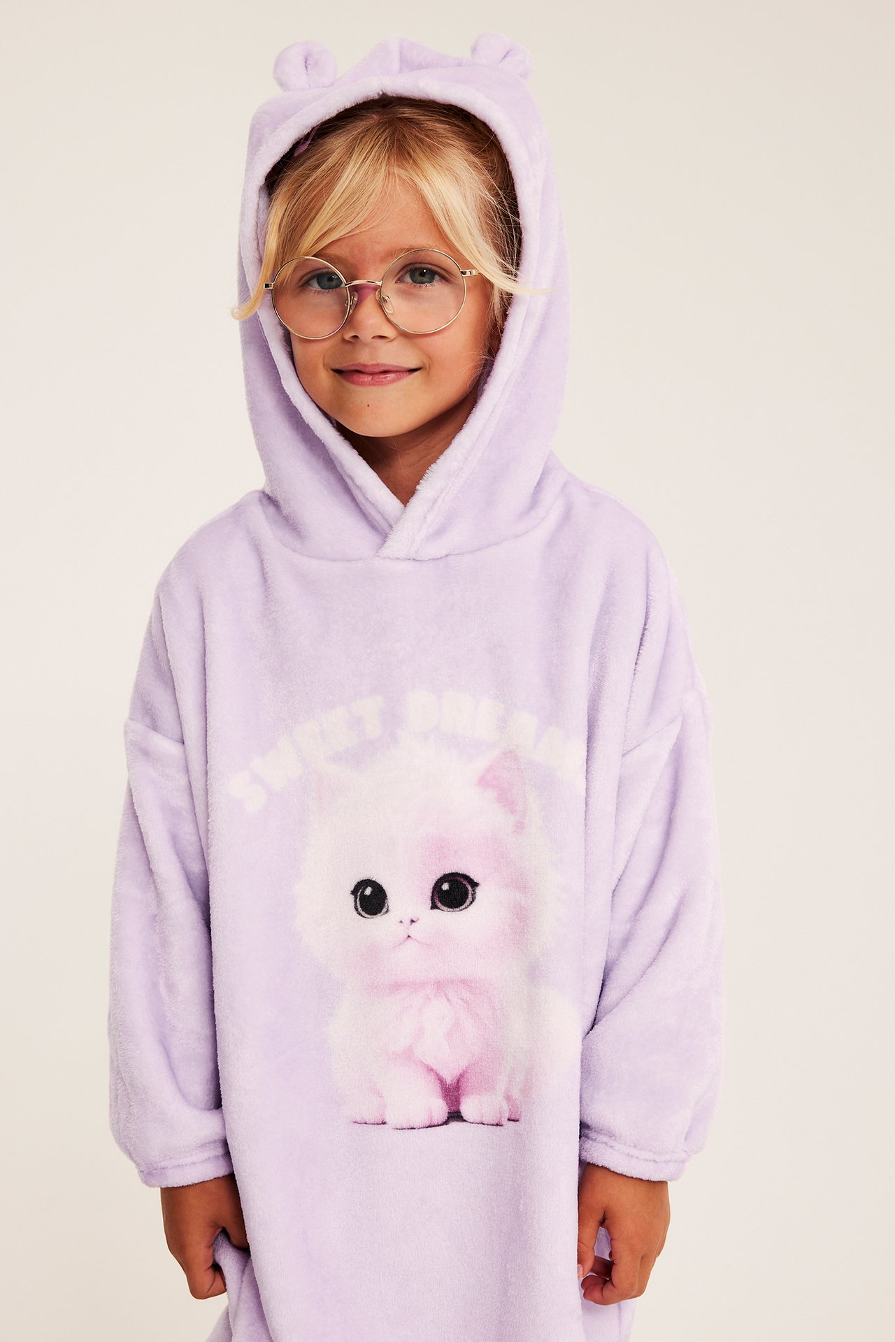 Barn framifrån i lila fluffig oversize-filt med huva, kattryck och runda glasögon.
