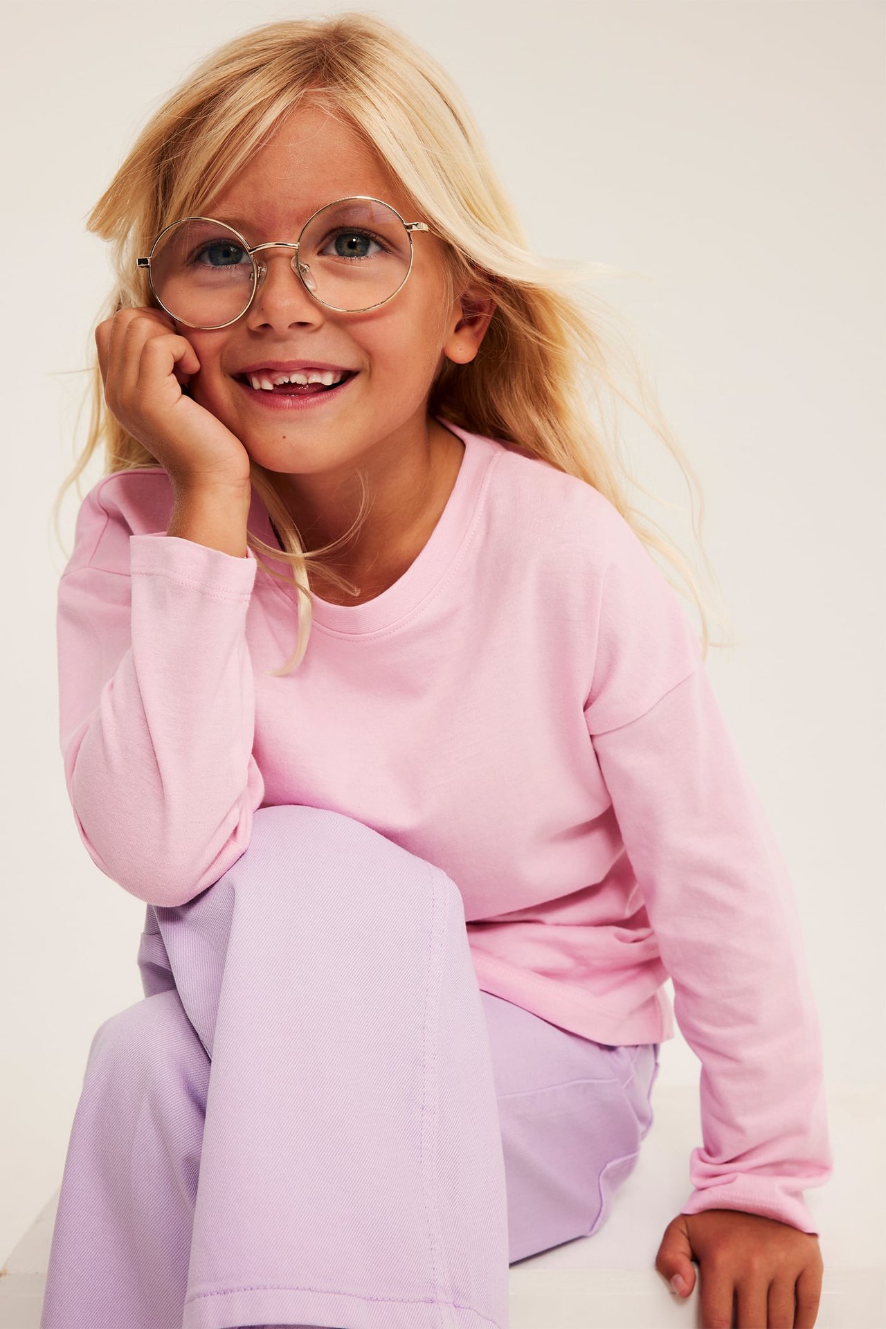 Framsidan av ett leende barn med rosa långärmad tröja, ljuslila byxor och runda glasögon.