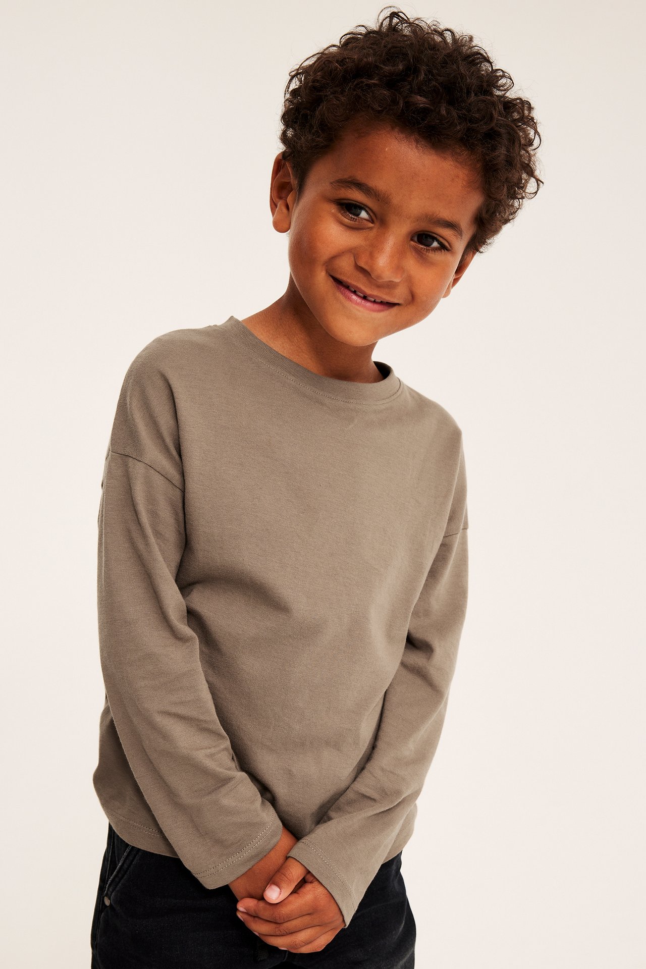 Framsida av ett barn i en långärmad brunbeige t-shirt och svarta byxor, leende.