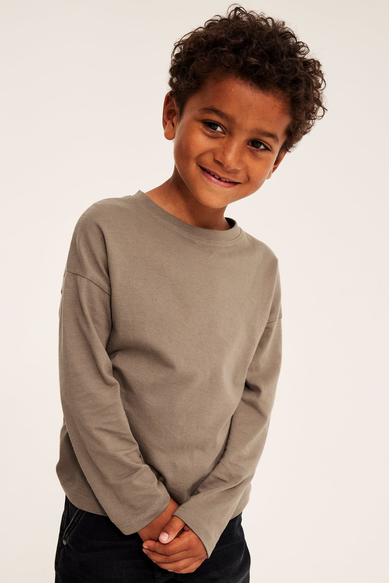 Framifrån av ett barn i en taupe avslappnad långärmad t-shirt och mörka byxor.