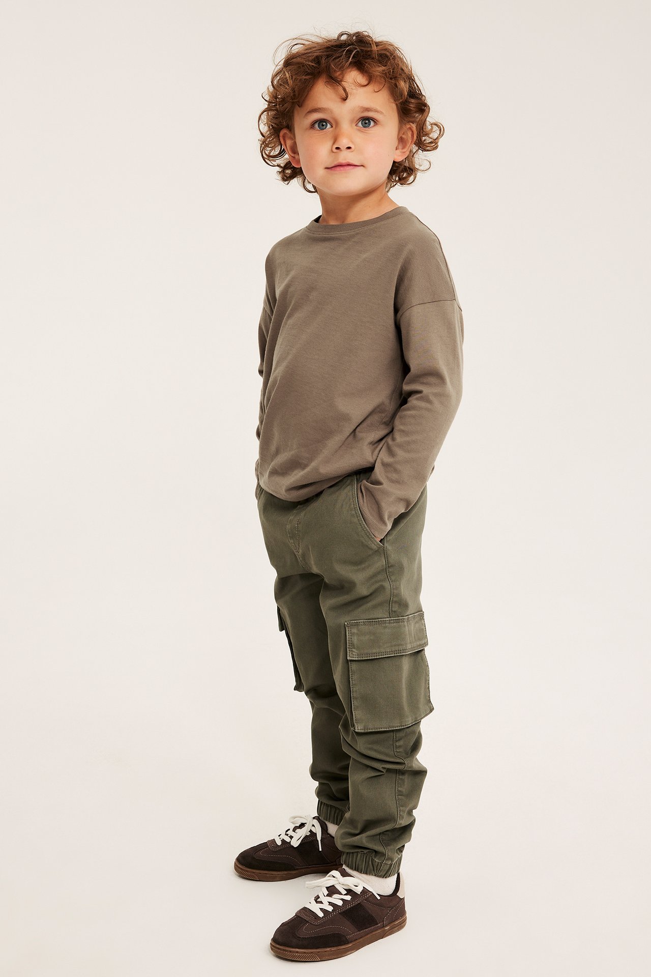 Dziecko z boku: brązowy oversize longsleeve, zielone spodnie cargo, ciemnobrązowe sneakersy.