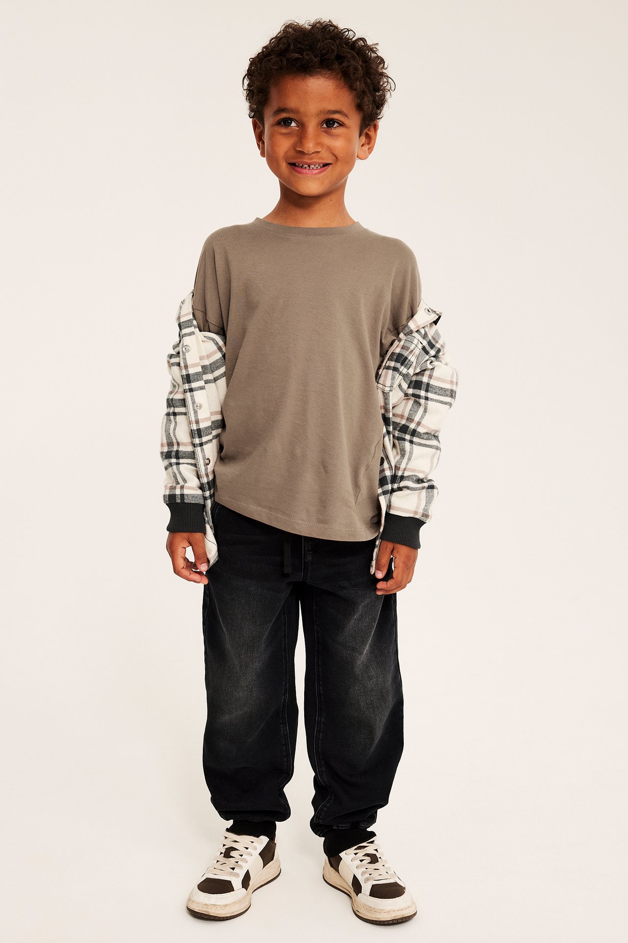 Framsida av barn i ljusbrun långärmad t-shirt, rutig skjorta, tvättade svarta jeans och sneakers.