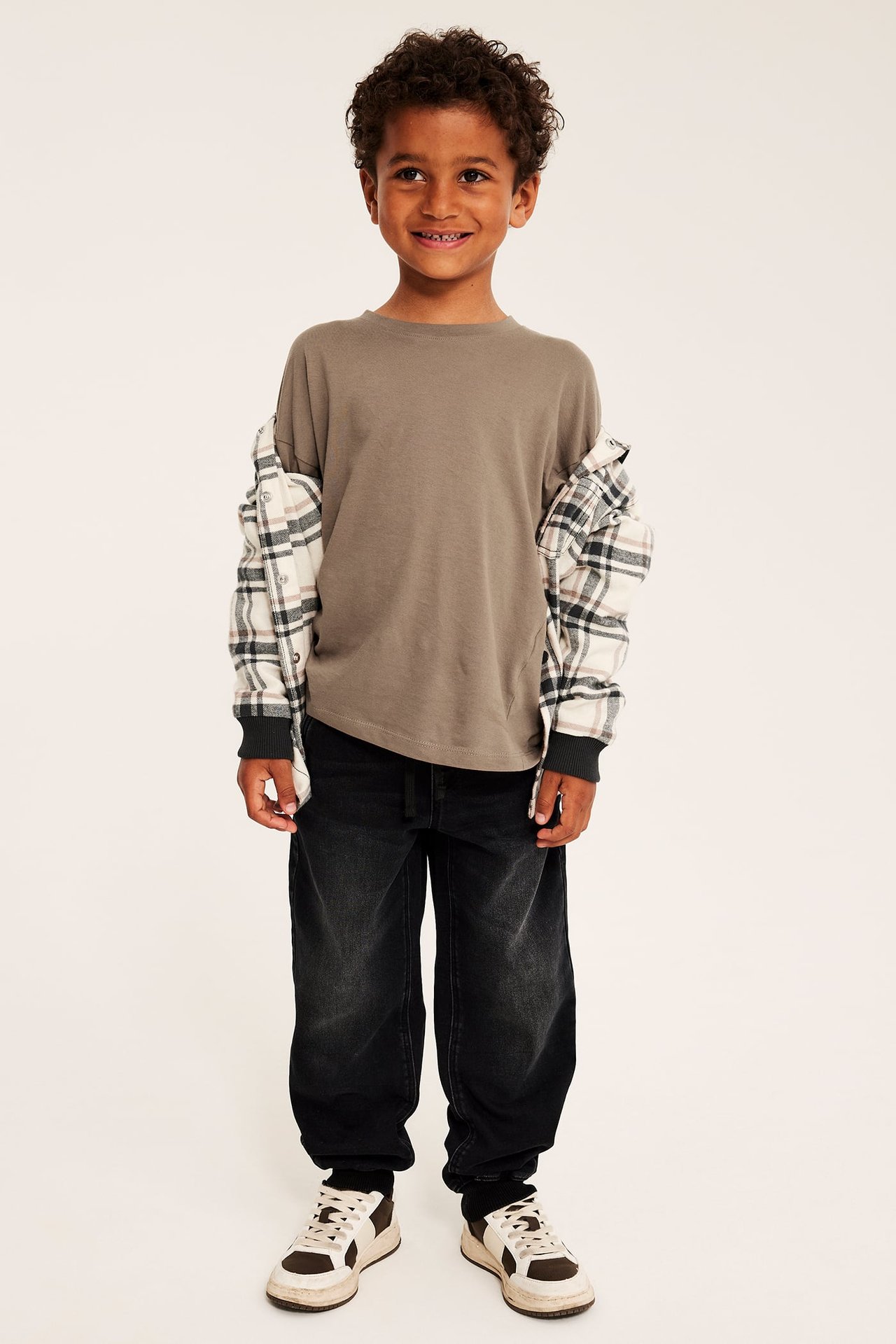 Framsida av barn i ljusbrun långärmad t-shirt, rutig skjorta, tvättade svarta jeans och sneakers.