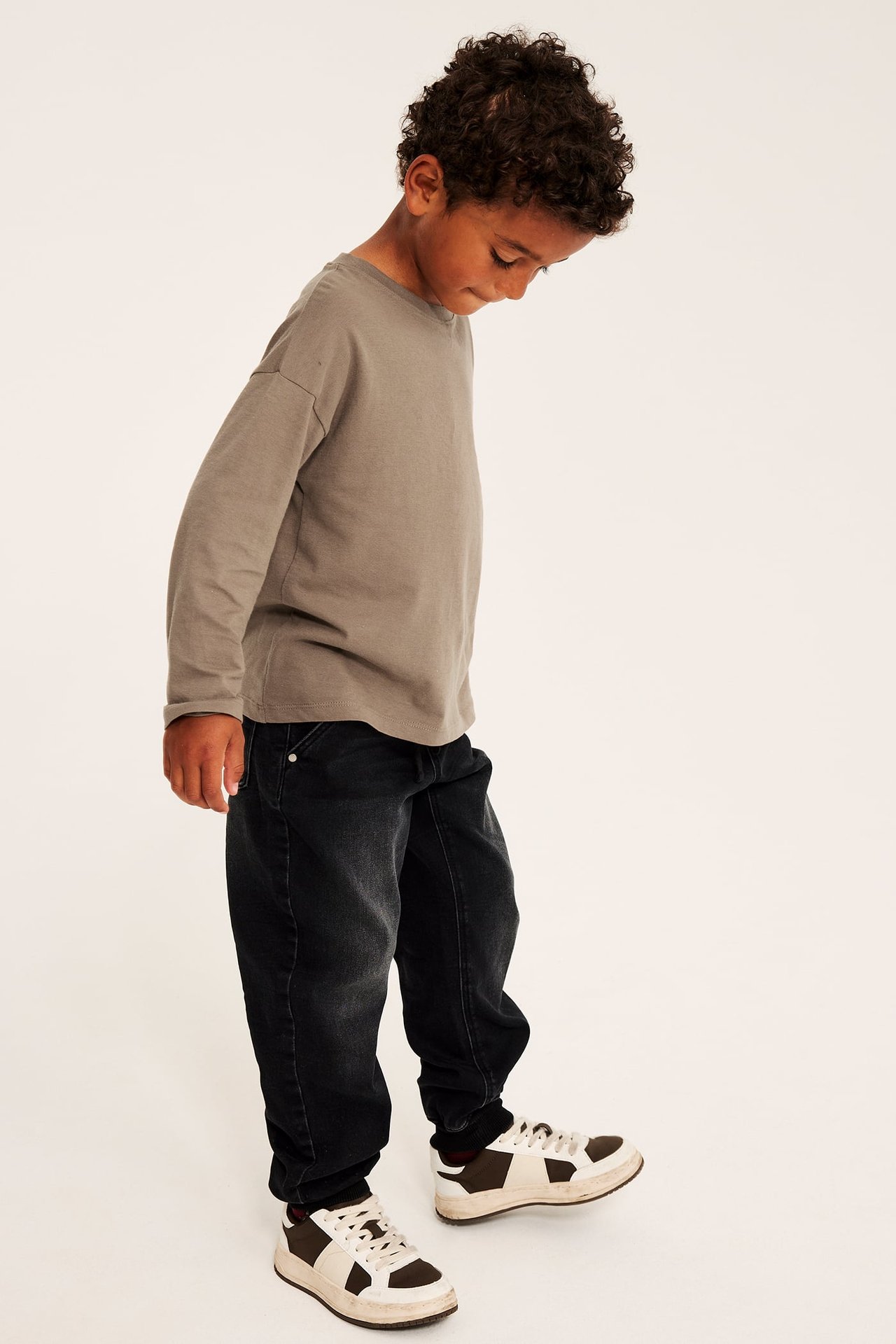 Lös grå långärmad t-shirt på ett barn, med svarta jeans och tvåfärgade sneakers, sidovy.