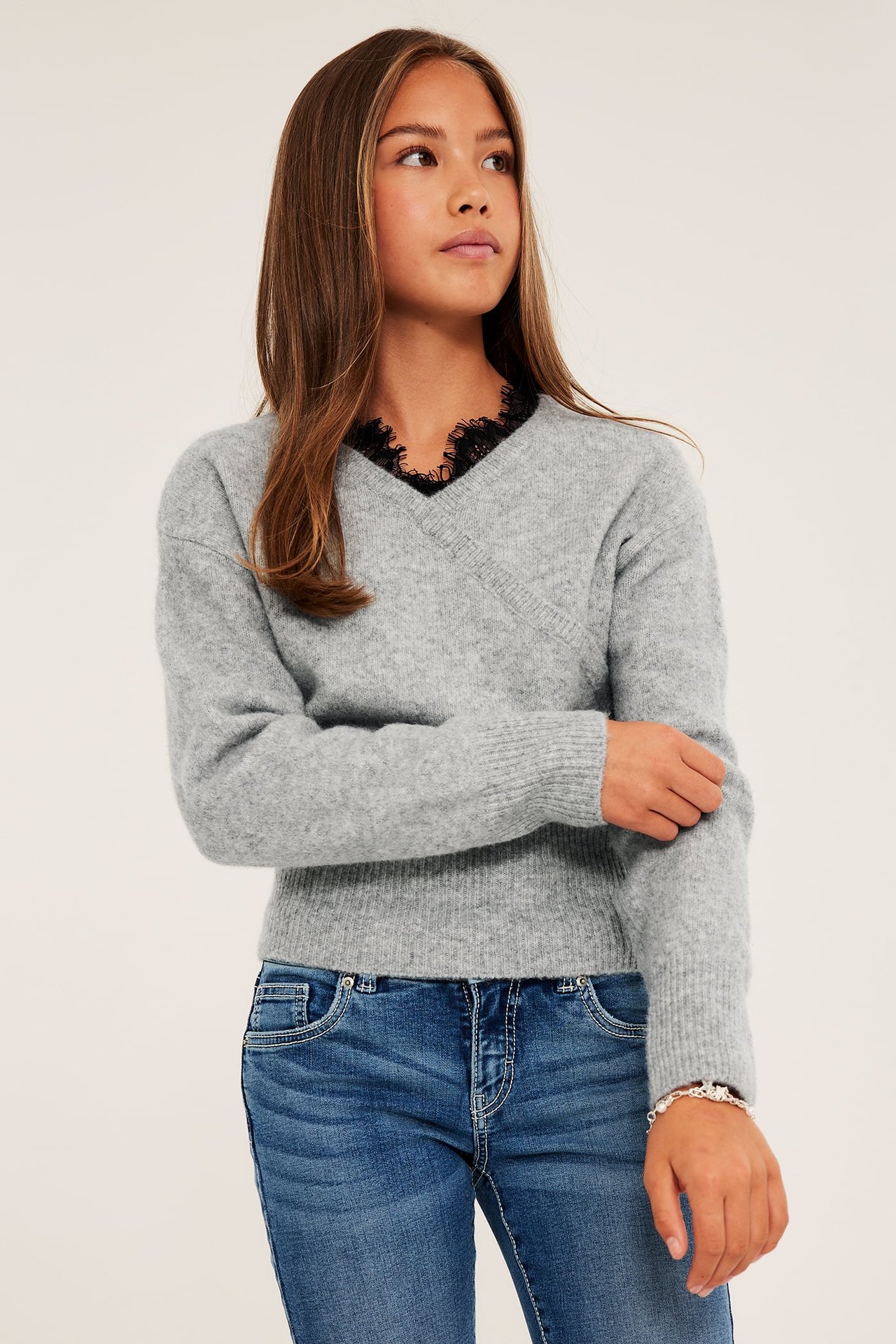 Grå omslagsgenser med V-hals og svart blondekant, blå jeans og et sølvarmbånd på et barn.